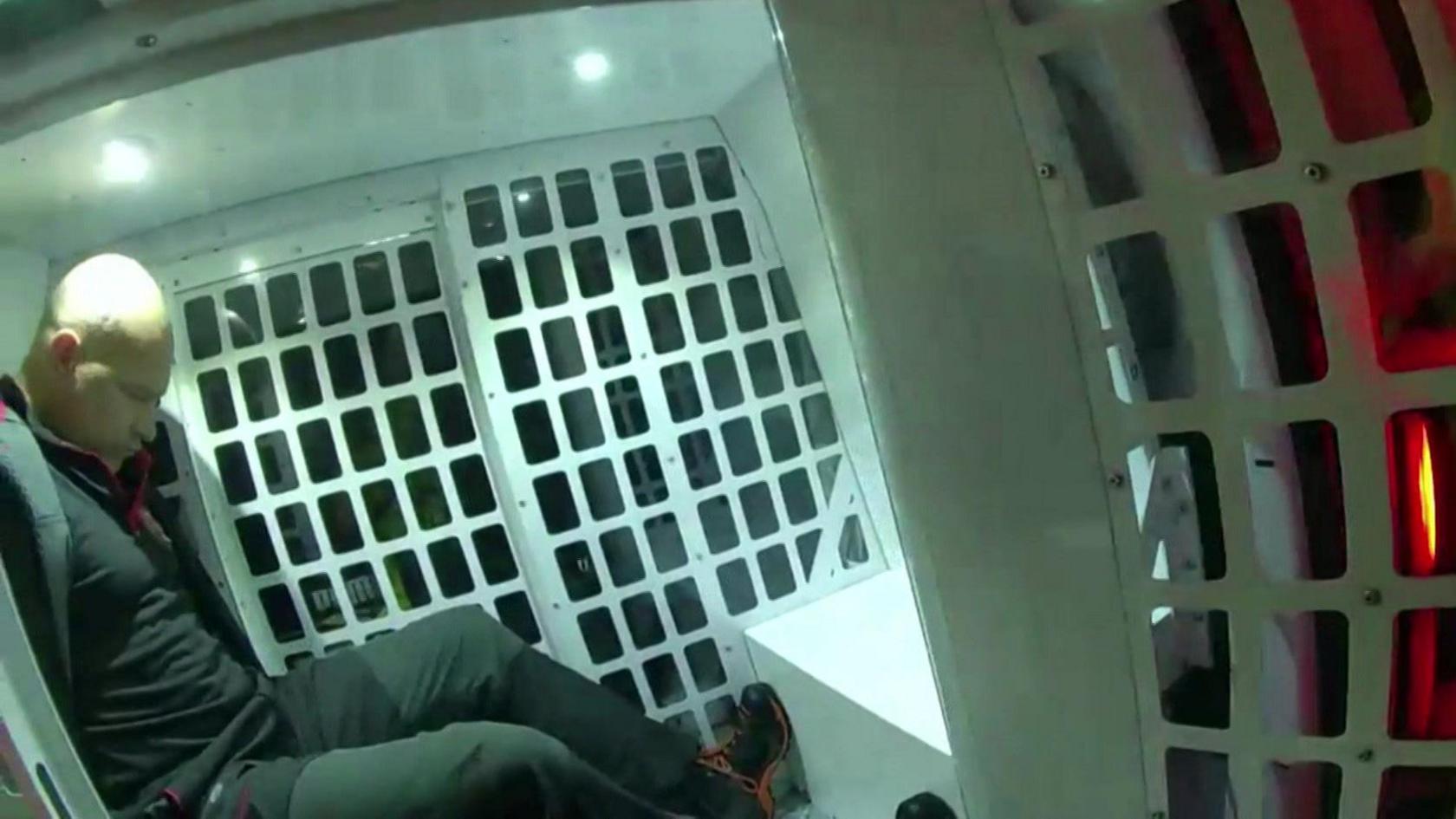 Police body camera footage of a man with his arms behind his back in a police vehicle. There's a white grille behind him and the back doors are open. he is dressed all in black. He has his head looking down at his feet. he is bald and clean shaven.