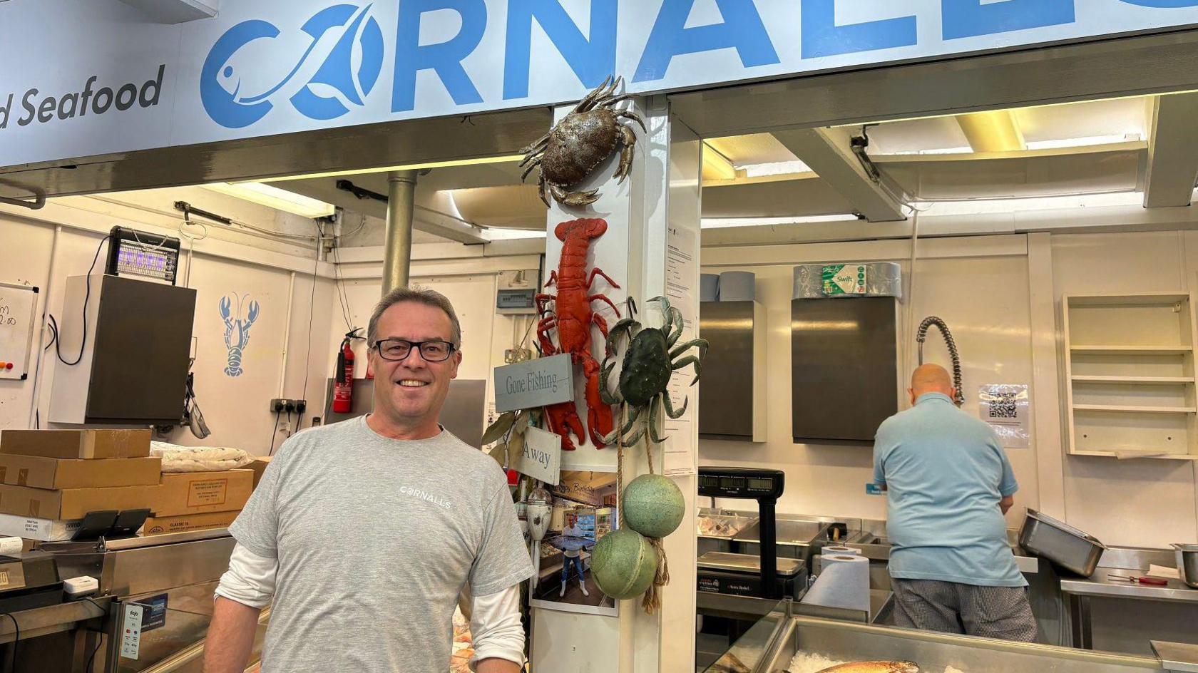 A man with short brown hair and black glasses is wearing a grey t-shirt with a white long sleeve underneath. He is standing in front of a fishmongers market staff that has a blue sign that reads "cornalls". On a pillar behind him, there are fake sea creatures, including a crab, and a lobster.