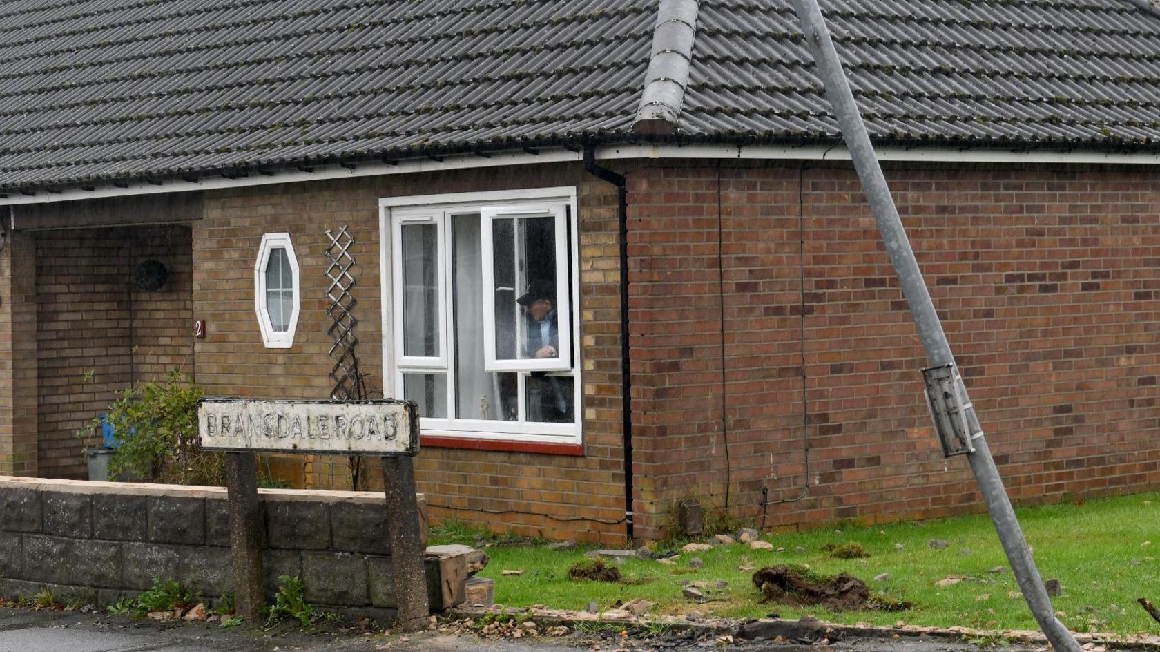 Street sign in front of a bungalow next to the remains of a garden wall with rubble on a lawn and a bent grey-coloured signpost 