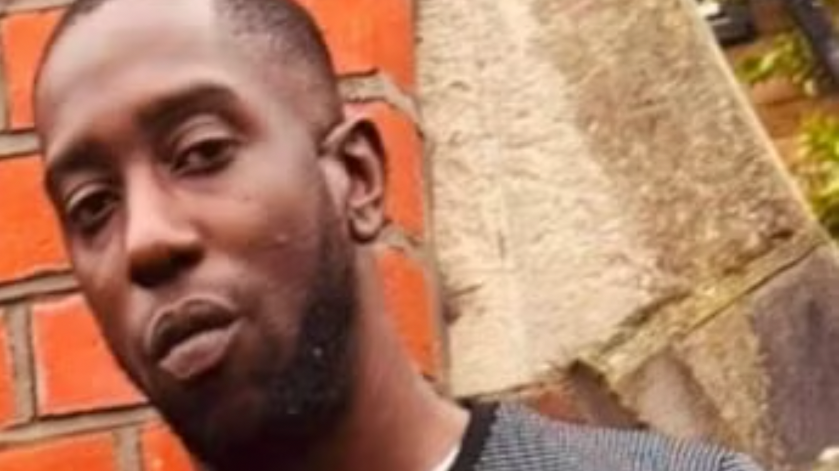 An image of Recorda Davey-Ann Clarke. He is looking towards the camera with some orange brickwork in a wall behind him. he has a buzzcut haircut and a short beard.