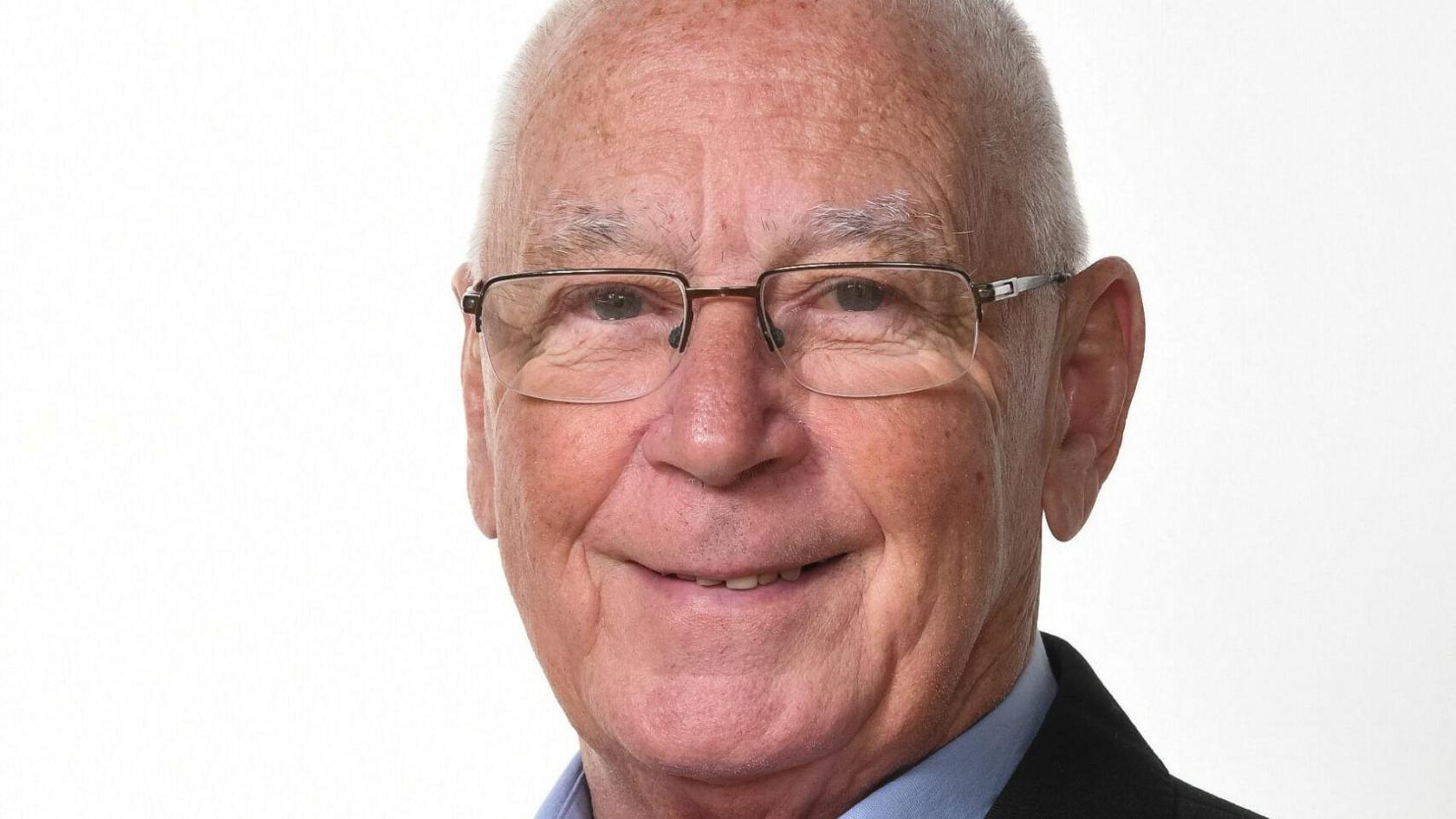 Sean Devlin smiling and looking into the camera. He is wearing glasses and a suit. He has a bald head. 