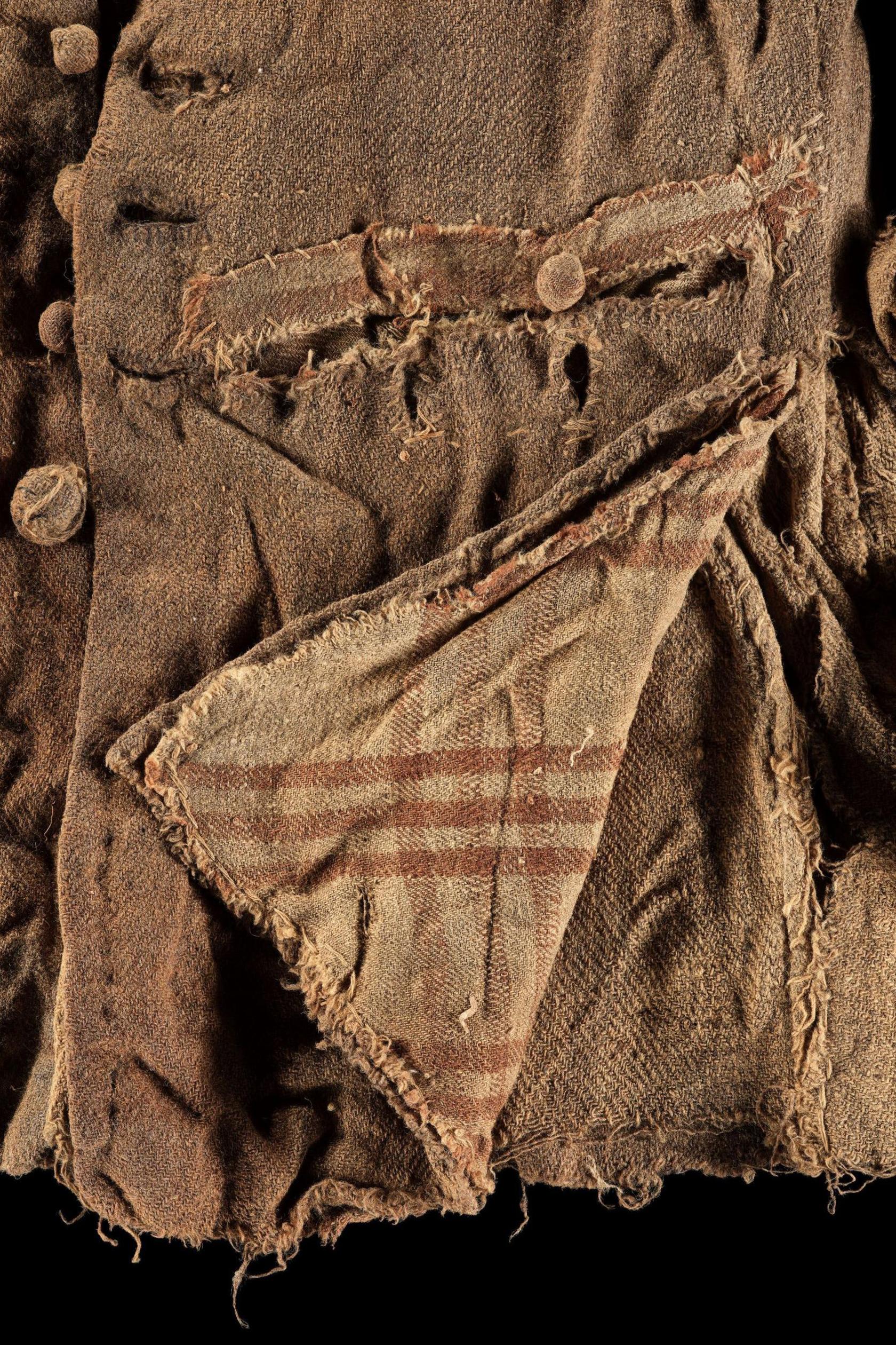 The jacket is brown and the fabric is crumpled. There are buttons on the jacket and part of the jacket has been folded over to show a tartan pattern.