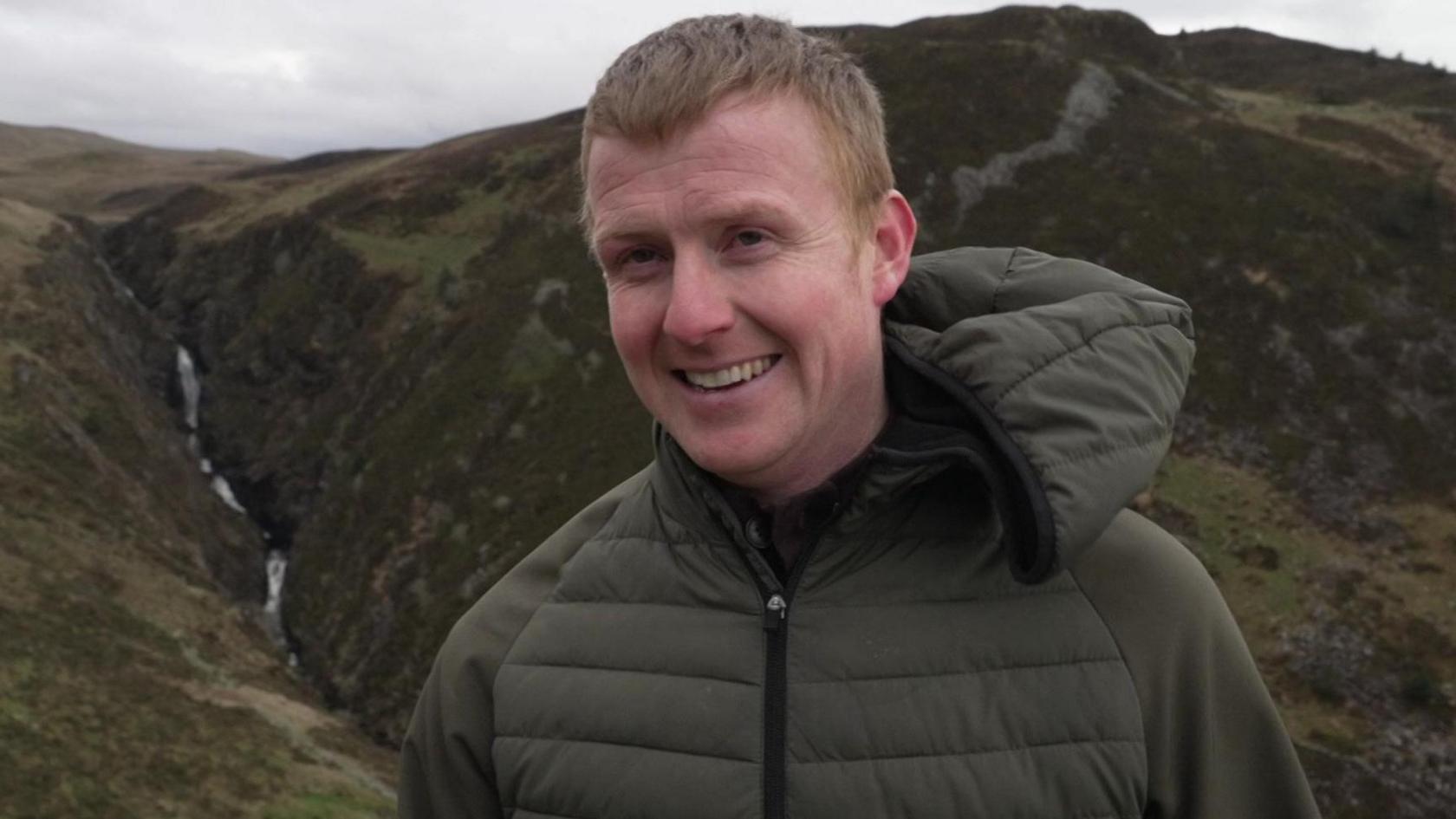 Llun Moi Dafydd gyda rhaeadr tu ôl iddo