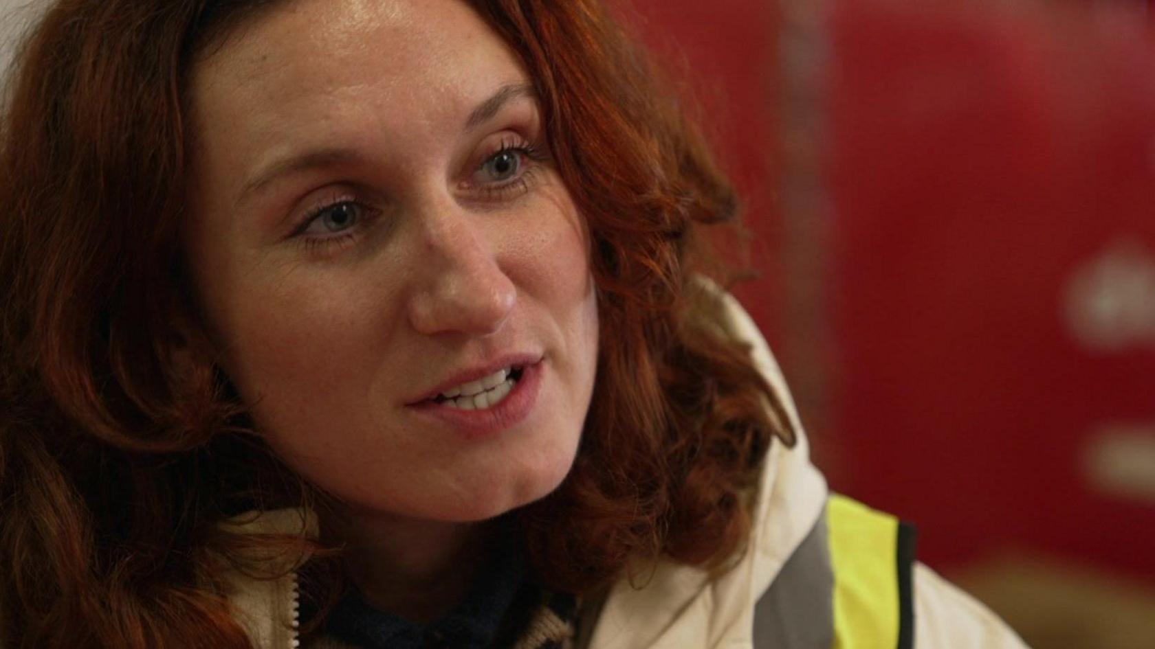 A close up of Bessie Carter's face. She is wearing a white coat and a high vis tabard and has long dark red hair. 