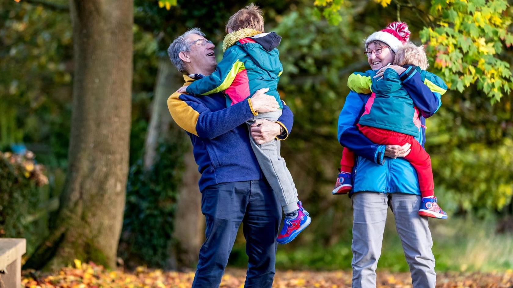 Judith and Martin Ellis stand outside, and hug two young children whose backs are to the camera. Against a backdrop of autumnal trees, Judith wears a red woolly hat, a blue coat and grey trousers. Martin wears a blue fleece with yellow shoulders and blue jeans.