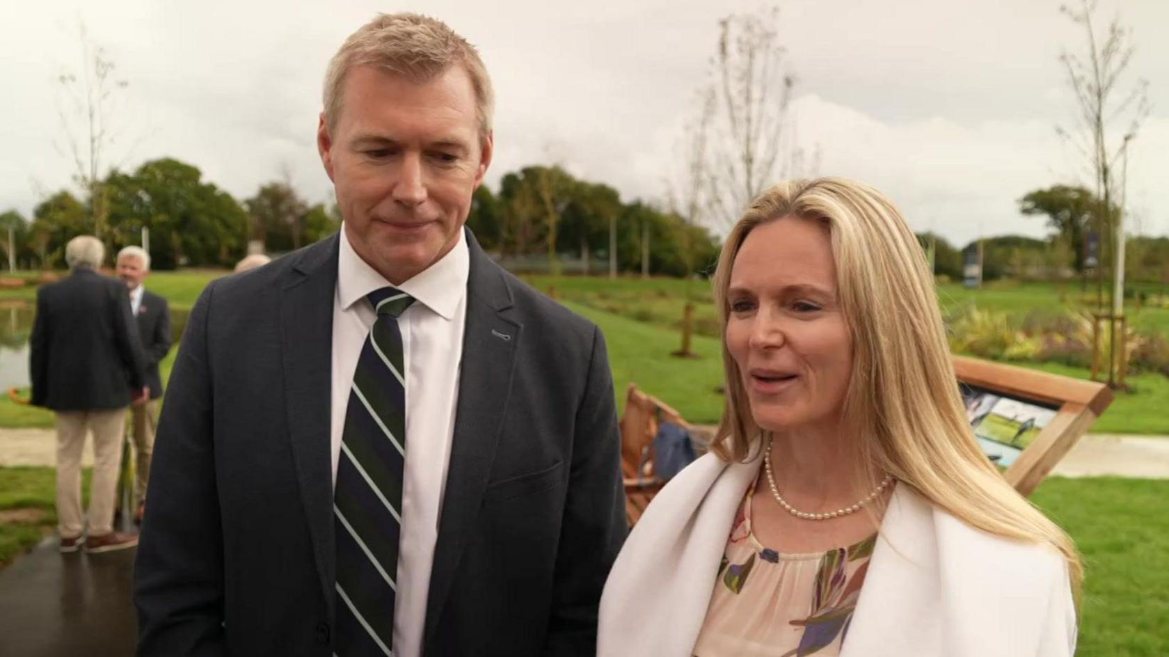 A tall caucasian man in a black suit with a striped tie stands in front of the memorial and beside his sister a shorter woman with long blonde hair wearing a floral dress and cream jacket. A park can be seen behind them. 