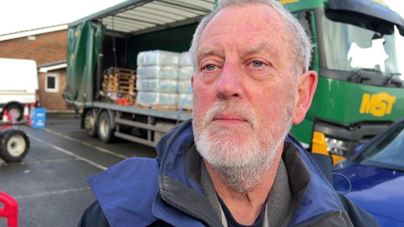 A man with short white hair and stubble wearing a blue coat. He is standing in front of a truck with bottled water in the back. 