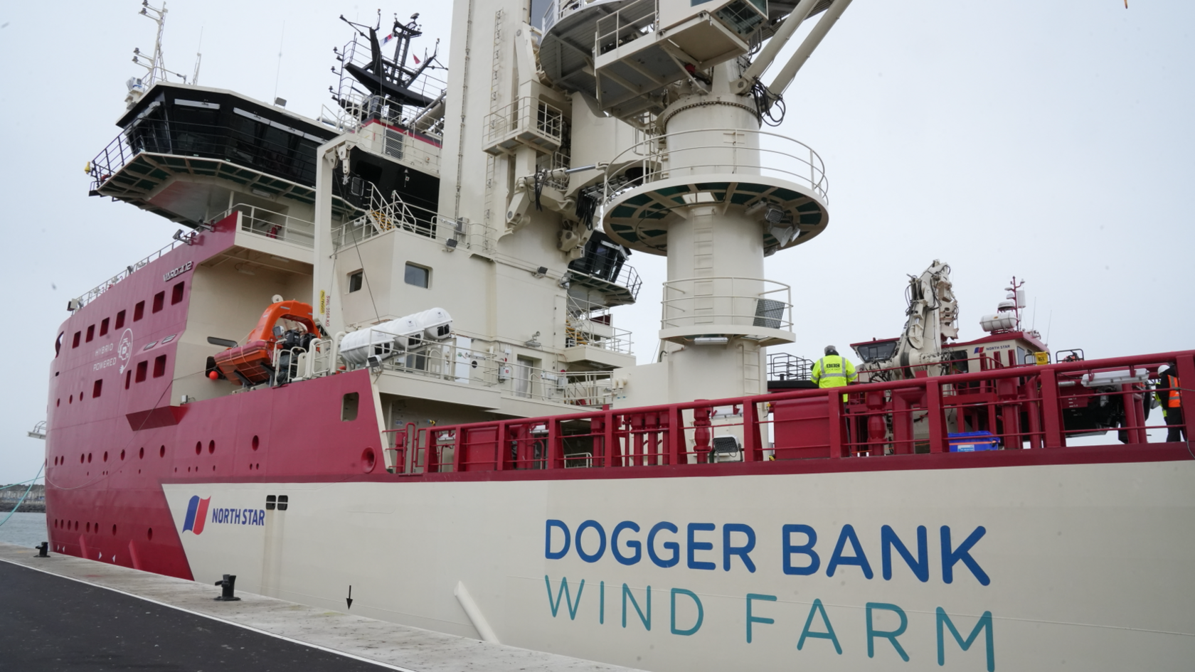 A large ship with the words Dogger Bank Wind Farm is docked at a port. It is red and white