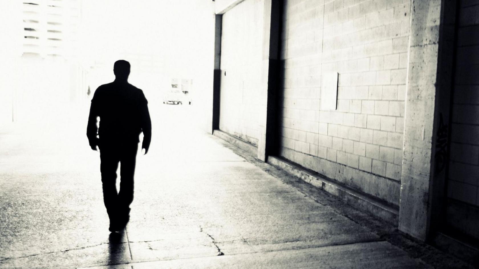 A black and white backlit silhouette of a man walking through an urban setting. The wall next to him is made of bricks.