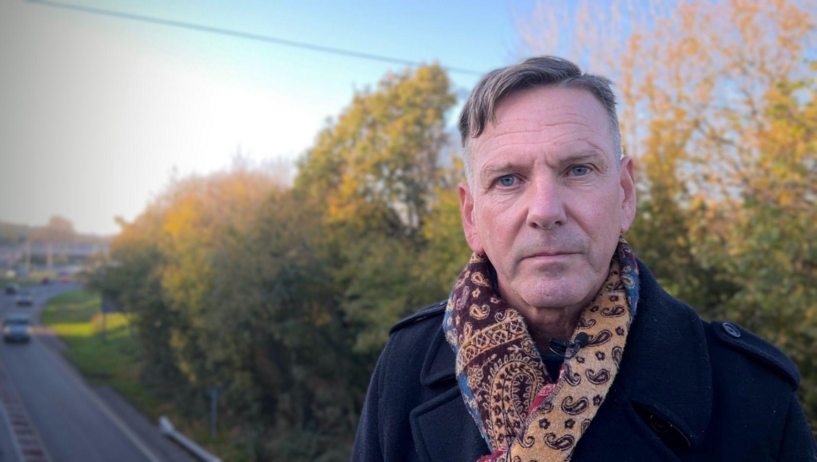 Richard Halliday, chairperson of Landkey Parish Council, wearing a patterned scarf and a navy blue coat, standing on a bridge overlooking the A361 North Devon Link Road