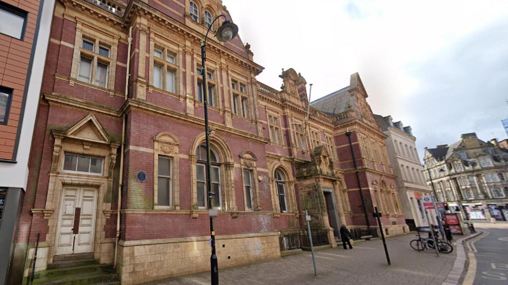 A view of a three-storey red-brick building as taken from below. There is a large expanse of pavement in front of it and buildings on either side. Other town buildings can be seen in the distance.