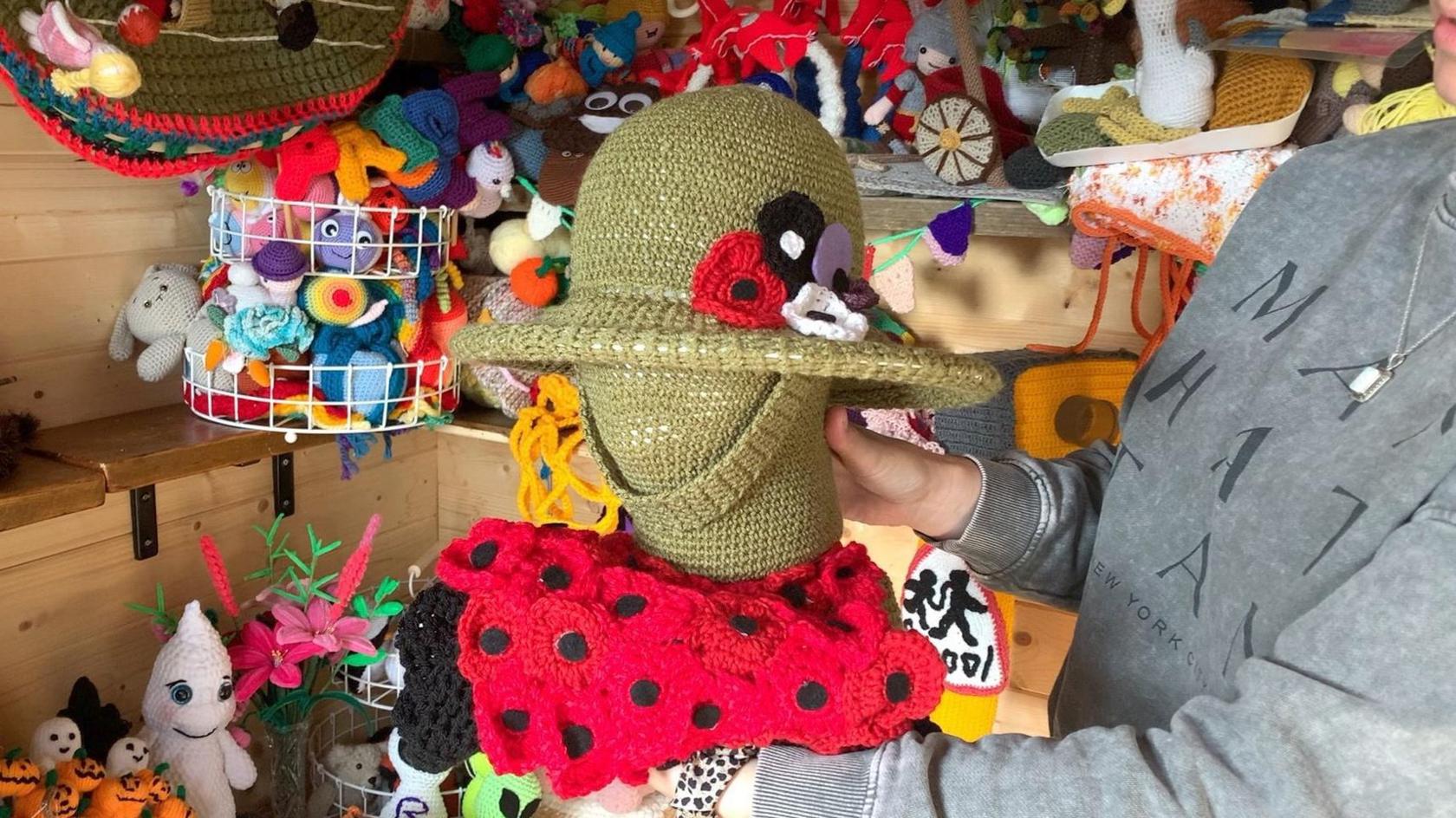 Alison Davis holds her Remembrance Day topper - a knitted green head and helmet, surrounded by poppies.  In the background are shelves full of more knitted toppers.