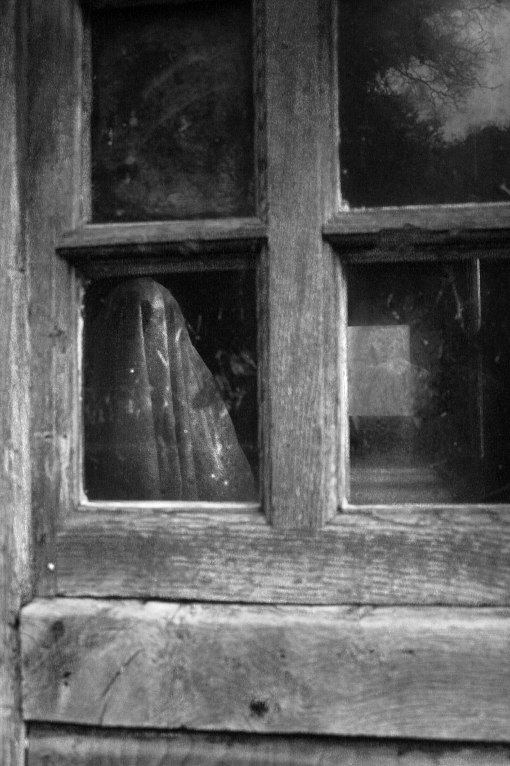 A black and white photograph of a window. Through the glass, a ghostly figure can be seen.