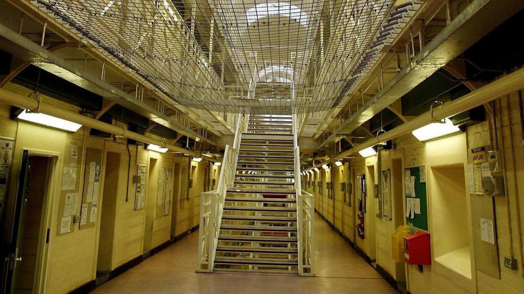 A prison showing cells either side with a central staircase going up to the next floor. The opening to the second floor is obscured by netting stretched across the gap.