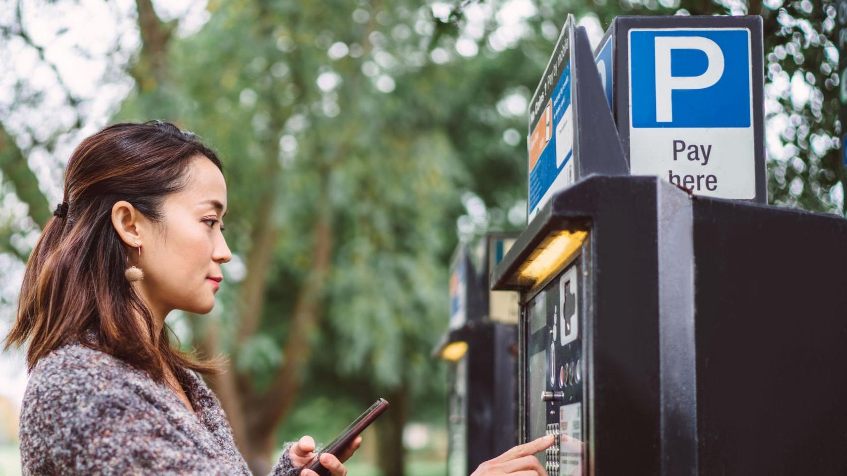 A young woman uses a parking payment machine in a city street. She has short brown hair tied back and a fluffy jumper