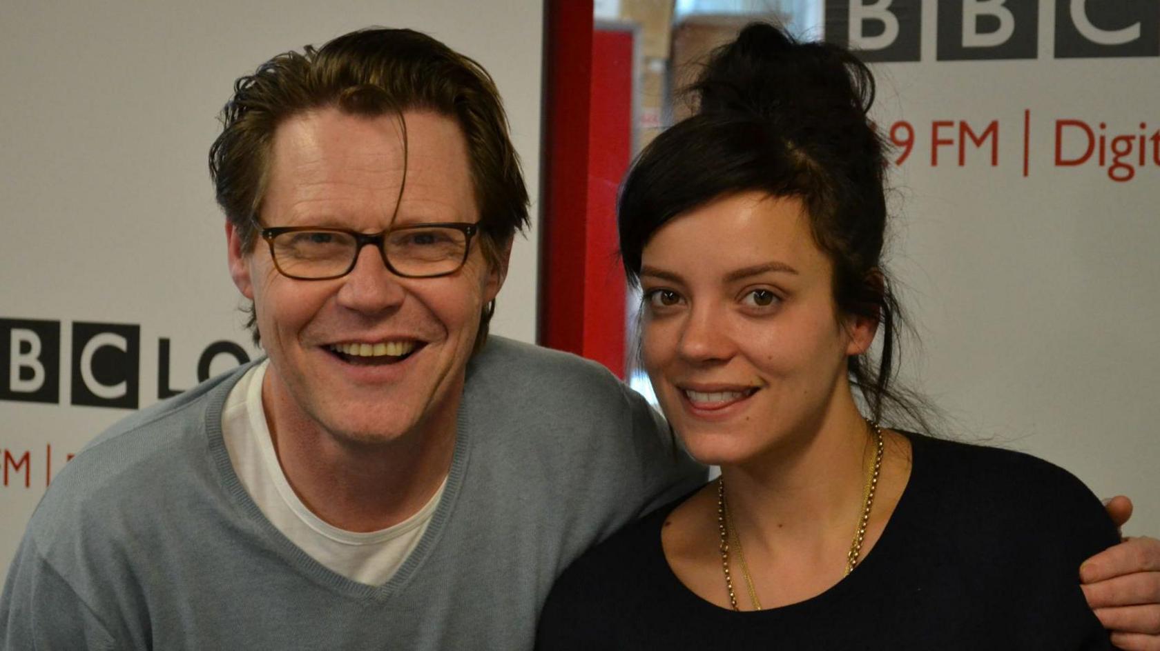 Robert Elms wearing a grey jumper over a white T-shirt with his arm around Lily Allen who is wearing a black top