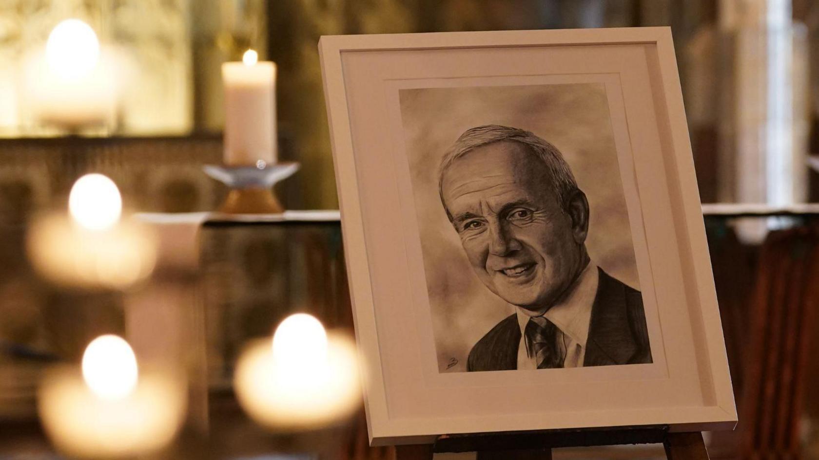 A portrait of Derek Underwood at Canterbury Cathedral, Kent, in a white frame. The portrait is on a stand surrounded by candles