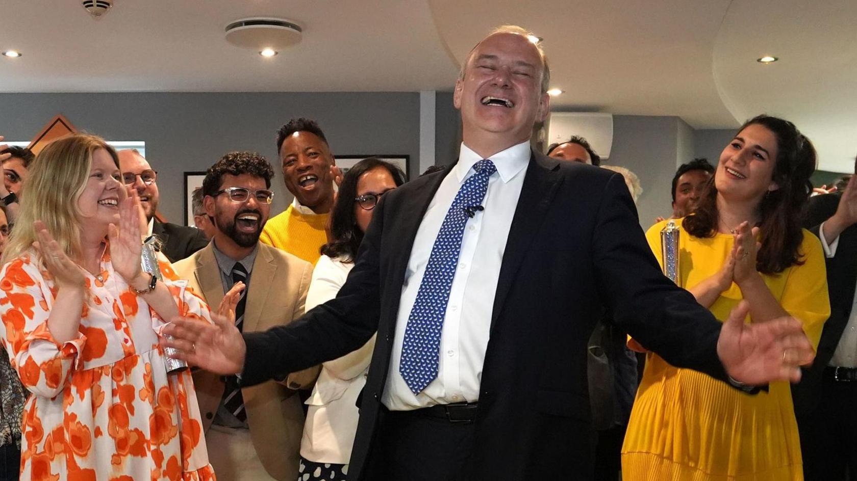 A very pleased looking Ed Davey with his arms apart celebrates with party supporters, some dressed in Lib Dem yellow