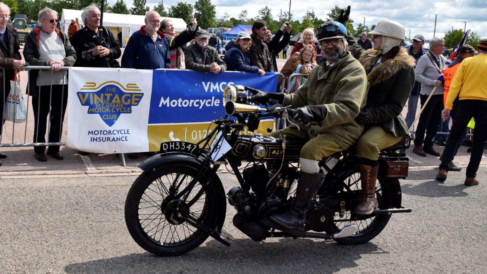 A man in old-school motorcycle clothing sits on a vintage bike with a woman on the back. They are both wearing helmets. Crowds of people are watching from the other side of a barrier.