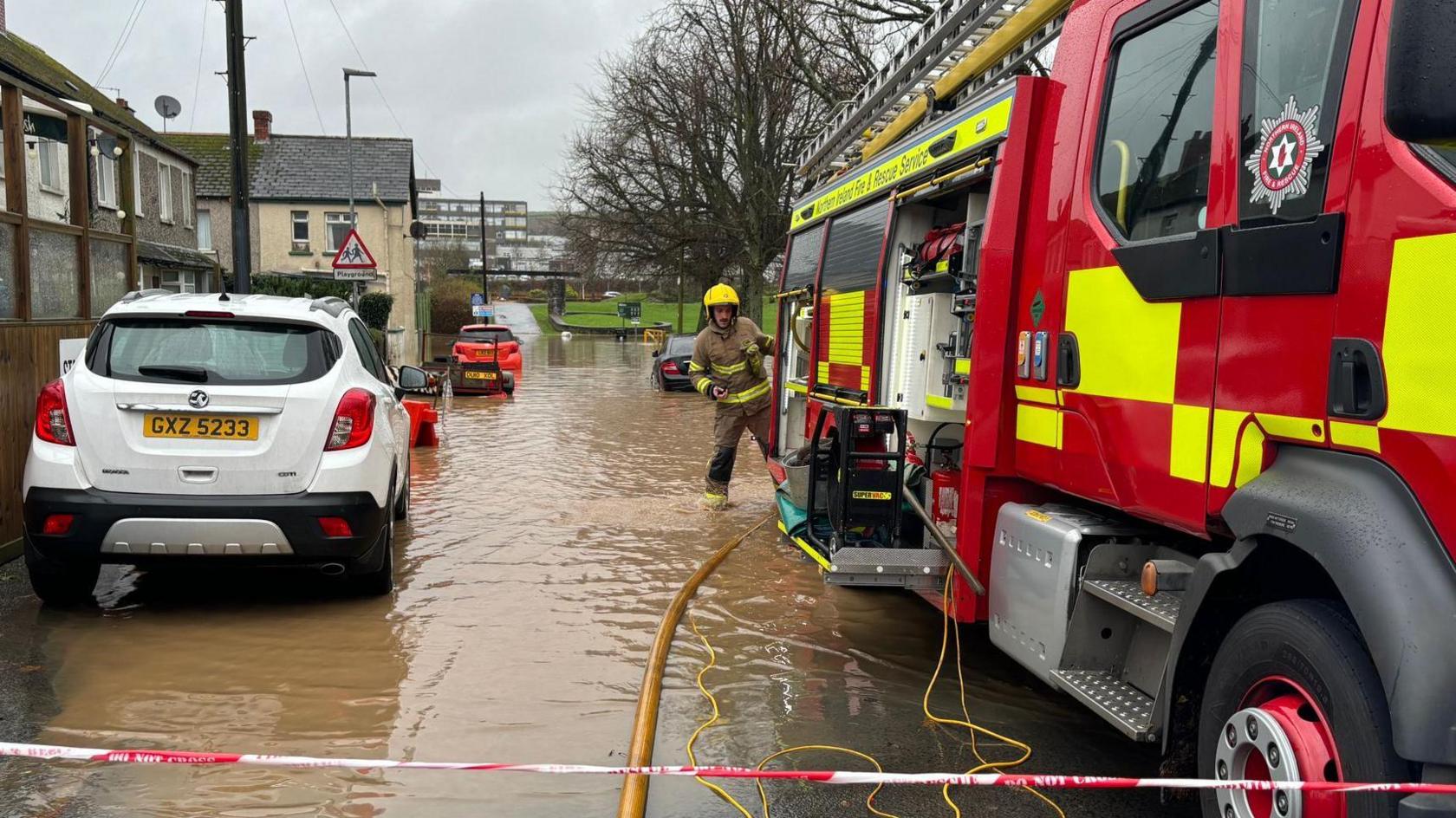 A picture of a flooded street - on the right is a fire engine, with a hose coming out of its rear. A firefighter stands at the back of the truck. On the ground is a foot or so of dirty brown water. On the left are parked cars and a row of terraced housing. 