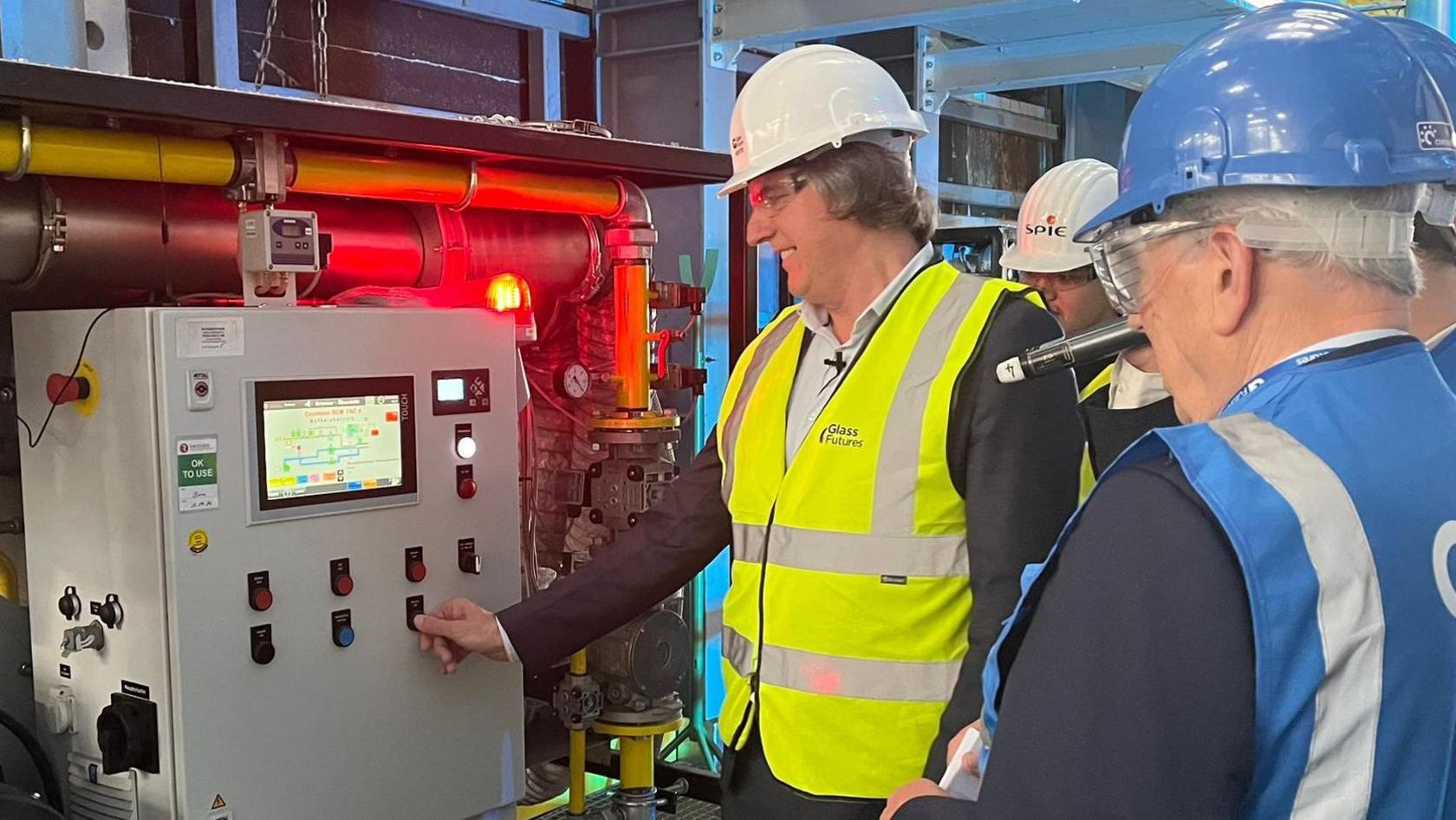 Steve Rotheram is wearing a black suit with a blue shirt and a high-visibility yellow jacket and white hard hat. He is pressing the button on a large grey metal box control system. He is stood alongside two people wearing blue high-visibility jackets and blue hard hats.