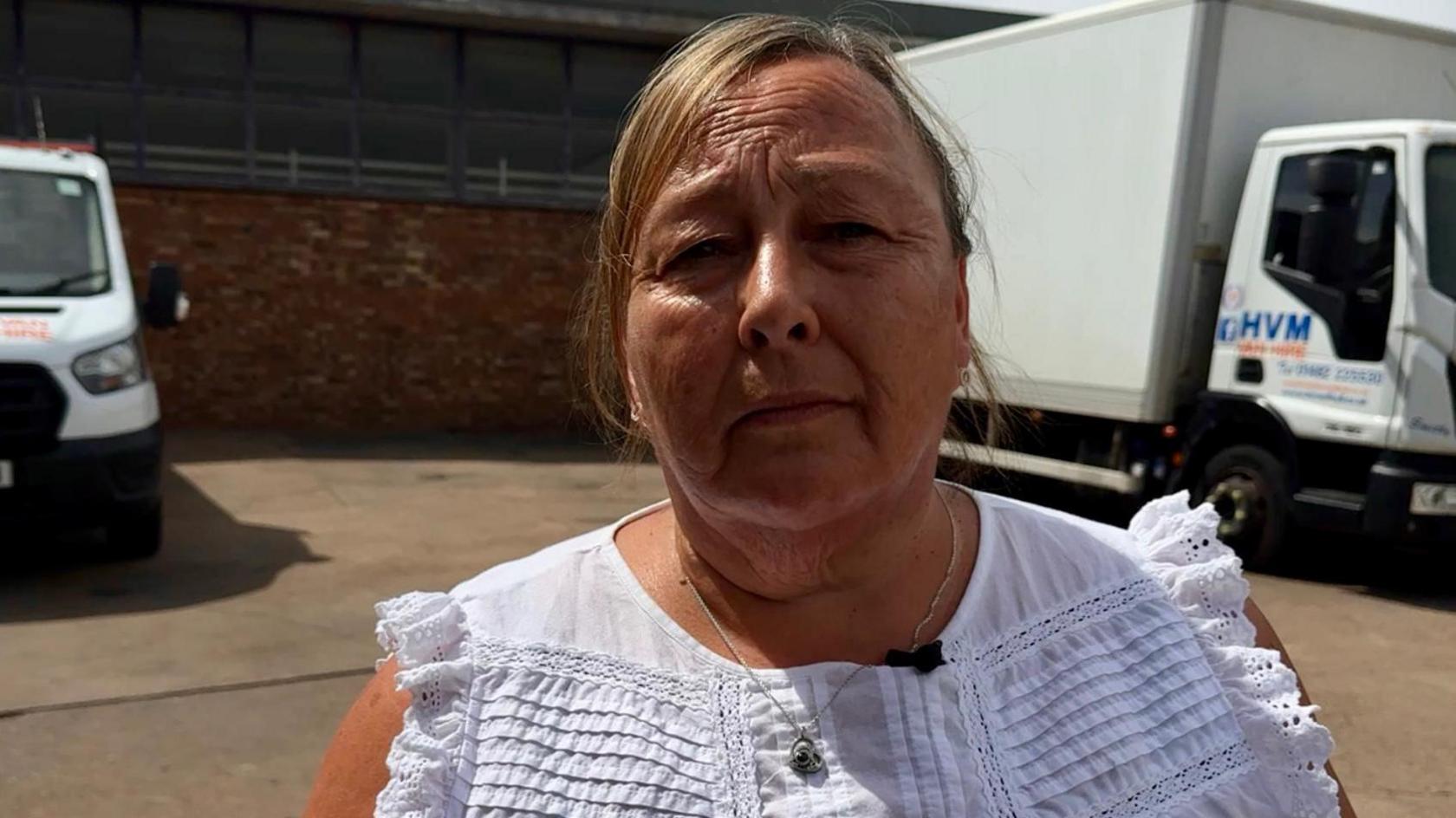 A woman with light brown hair and a white top looks seriously into the camera. Two white vans are visible in the background.