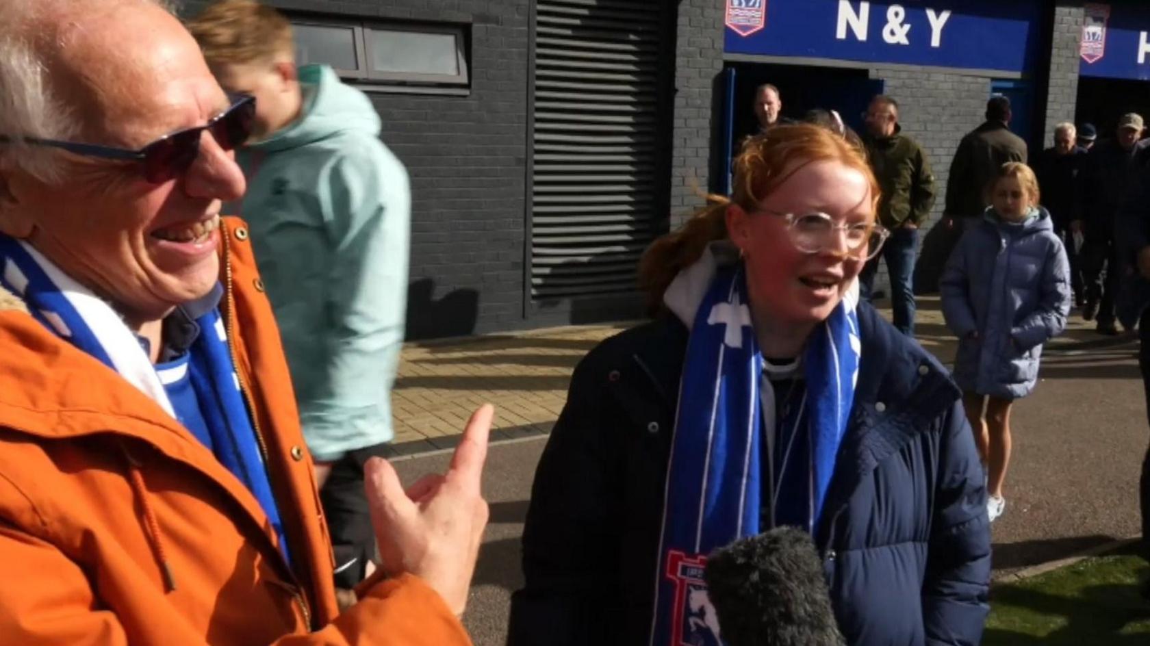 An older man in an orange jacket and Ipswich Town's blue underneath, stands facing a reporter who is off camera. He is pointing to a young woman with red hair beside him, who is in a navy coat.