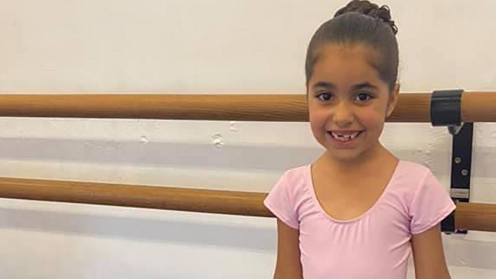 Alice with her brown hair in a bun dressed in a pink leotard in a dance studio. She is smiling.