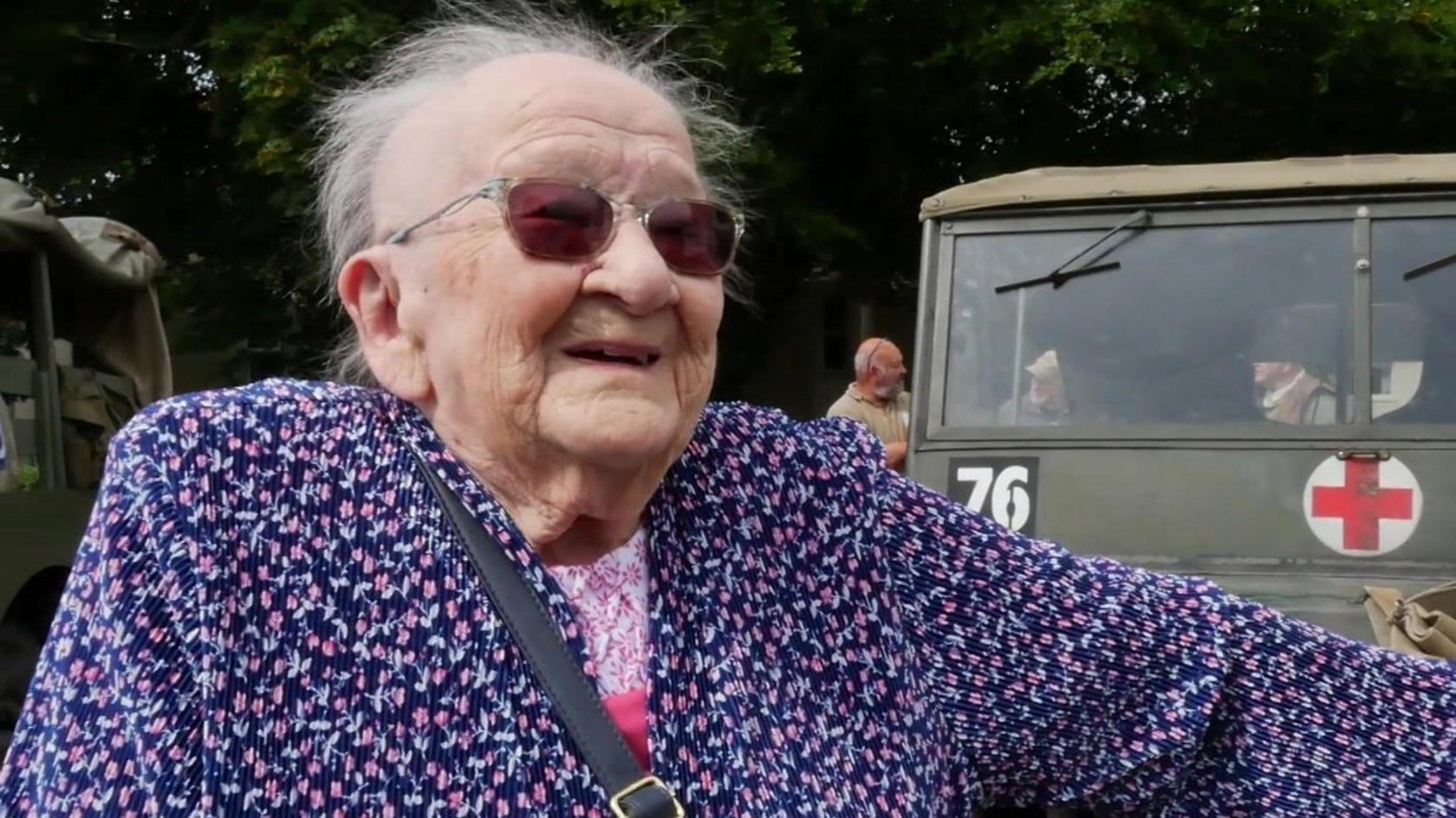 Kay White smiling in front of a green vintage World War Two vehicle which has the number 76 written on it and a red cross.