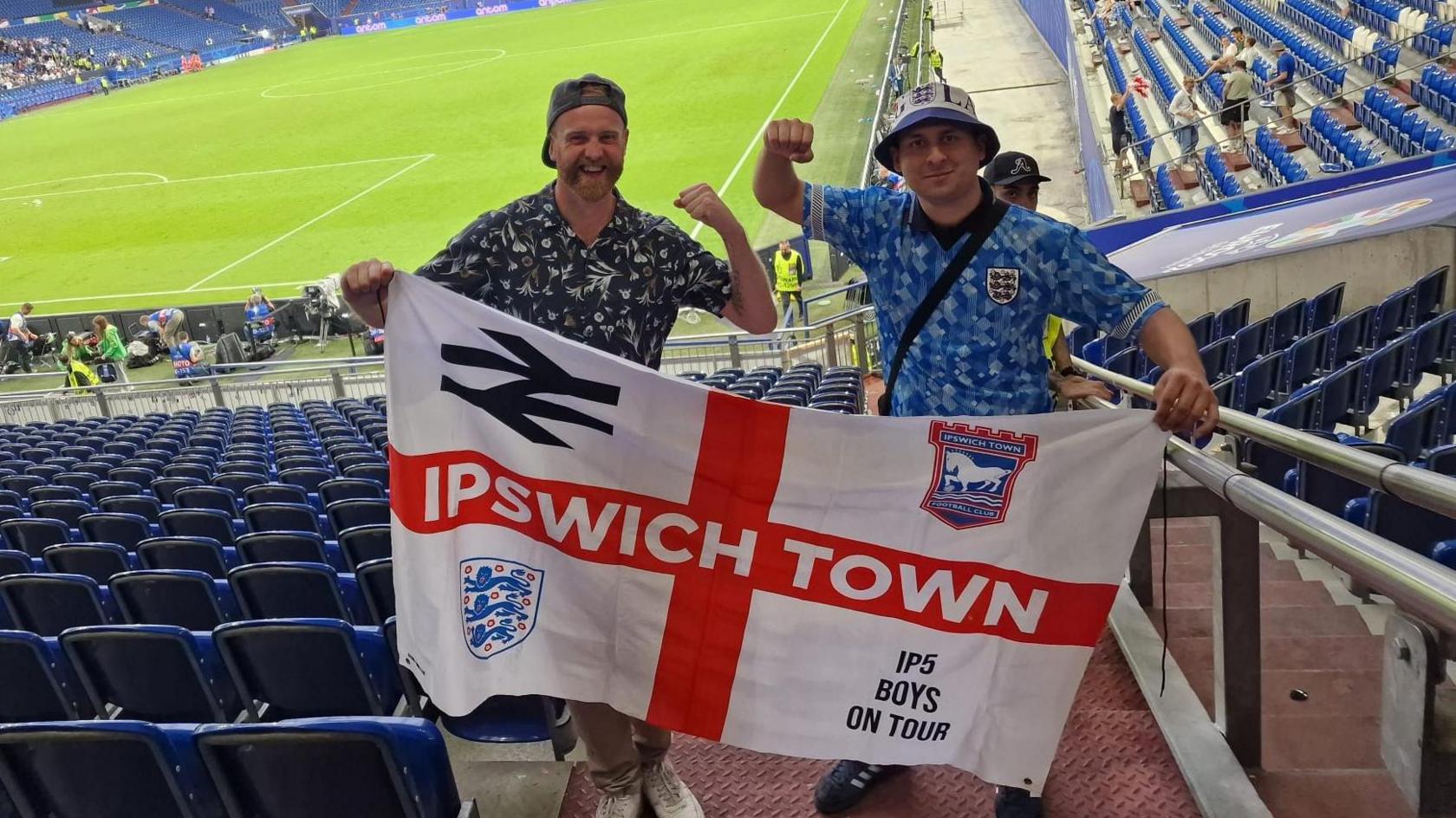 Karim Goodchild stands in an empty stadium with an empty football pitch behind them, he is dressed in a blue England shirt and is holding an Ipswich Town flag with another male