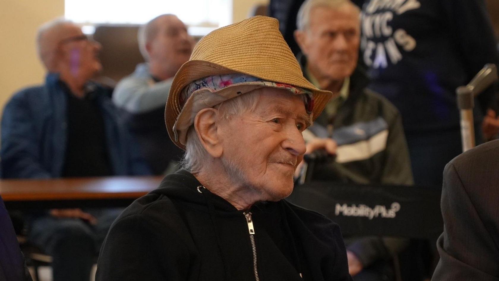 Ron "Ginger" Parsons looking to the right of the camera, watching a performance at his 103rd birthday party. He is wearing a straw hat over the top of a floral bucket hat. He has a black zipped up hoodie on. Behind him are three men also sat in the hall watching the performance. 