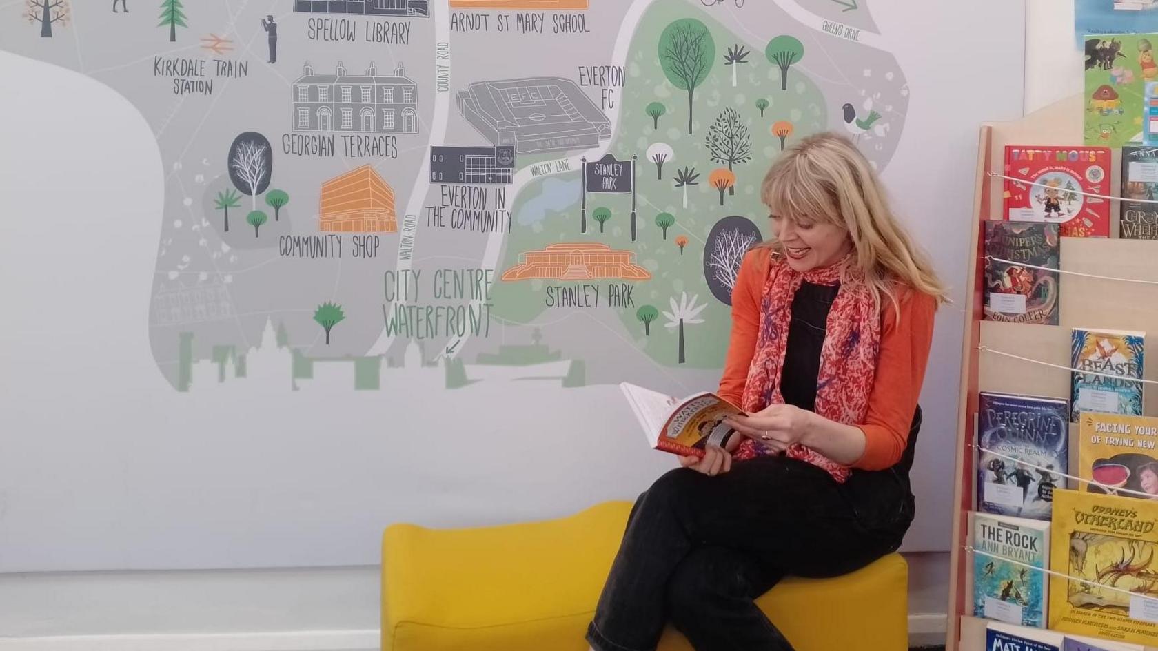 Artist Dora Colquhoun with long blond hair wearing an orange cardigan and black trousers and top sits on a yellow seat reading a book with a book case filled with books to the left. She sits in front of a wall with a map of Liverpool painted by an artist on the grey wall.