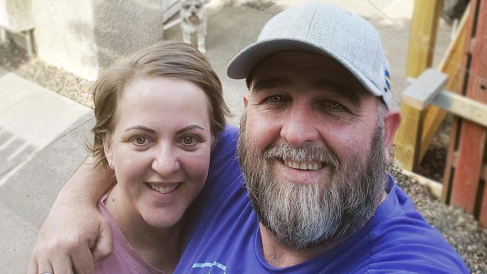 A man with a longer beard is standing in front of a house with his arm around a woman with short blond hair. She is wearing a pink top, while he is dressed in a dark-blue t-shirt and a grey cap. There is a schnauzer dog visible behind the couple.