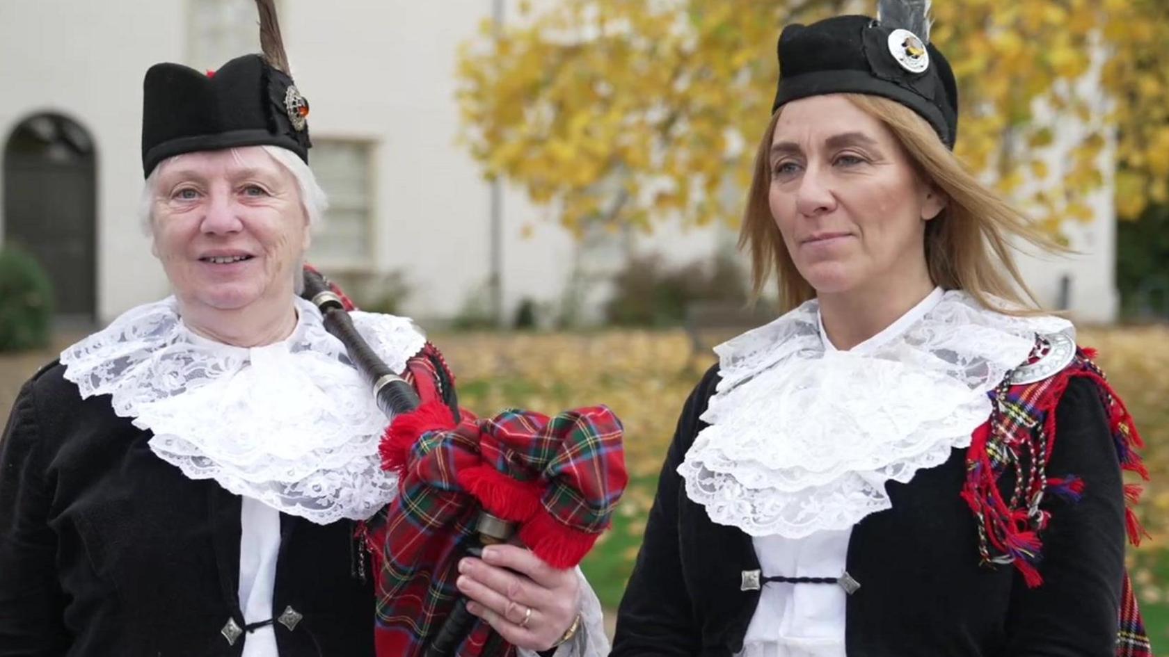 Sheila Hatcher and Denise Morrison standing outside in their uniforms, consisting of a black velvet jacket, red tartan bagpipes, white lace collars and black hats. A tree is visible in the background