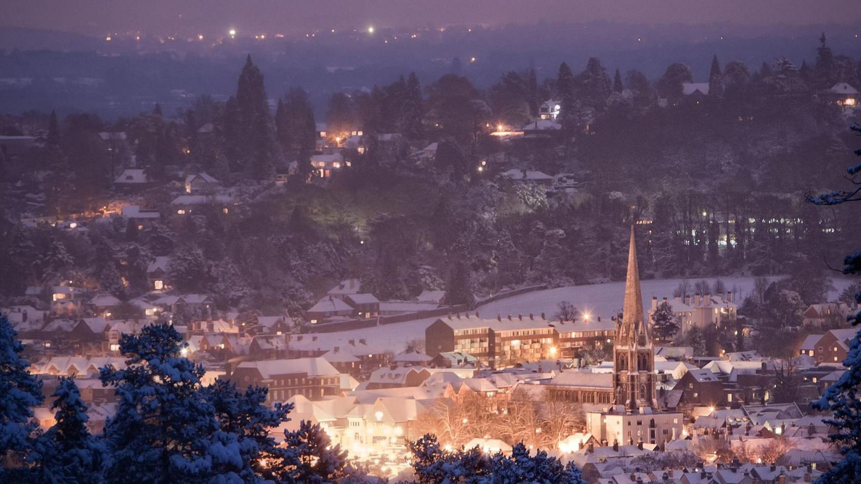 A snowy Dorking scene at night time, with trees in the foreground topped with snow, houses with white roofs and a church spire seen in the middle. In the distance more lights are seen on hills and among trees