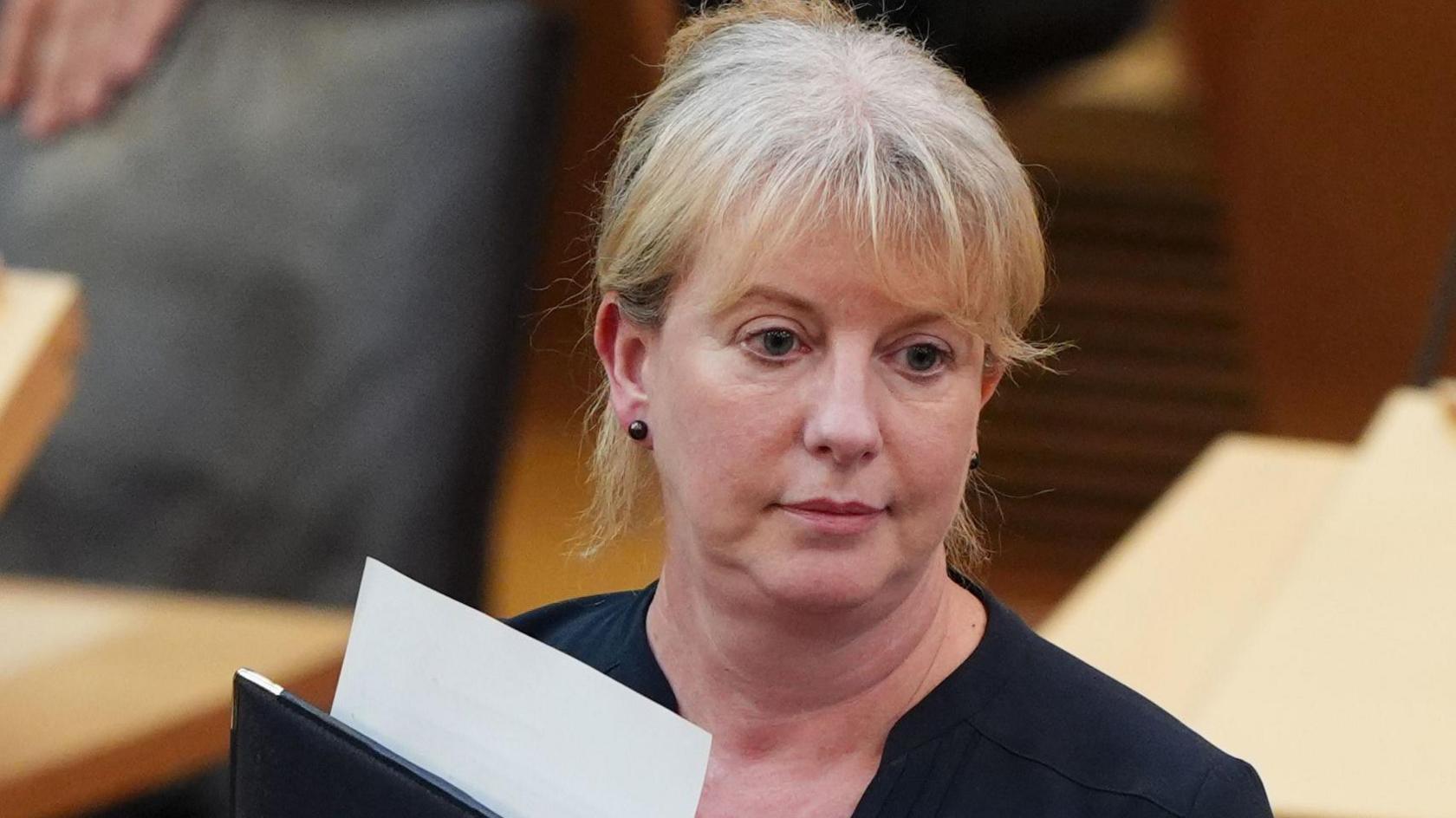 Shona Robison pictured in the Scottish parliament's main debating chamber. She has a blank expressions and appears deep in thought. Ms Robison is wearing a black dress and holding a black folder with a white sheet of paper protruding from the top