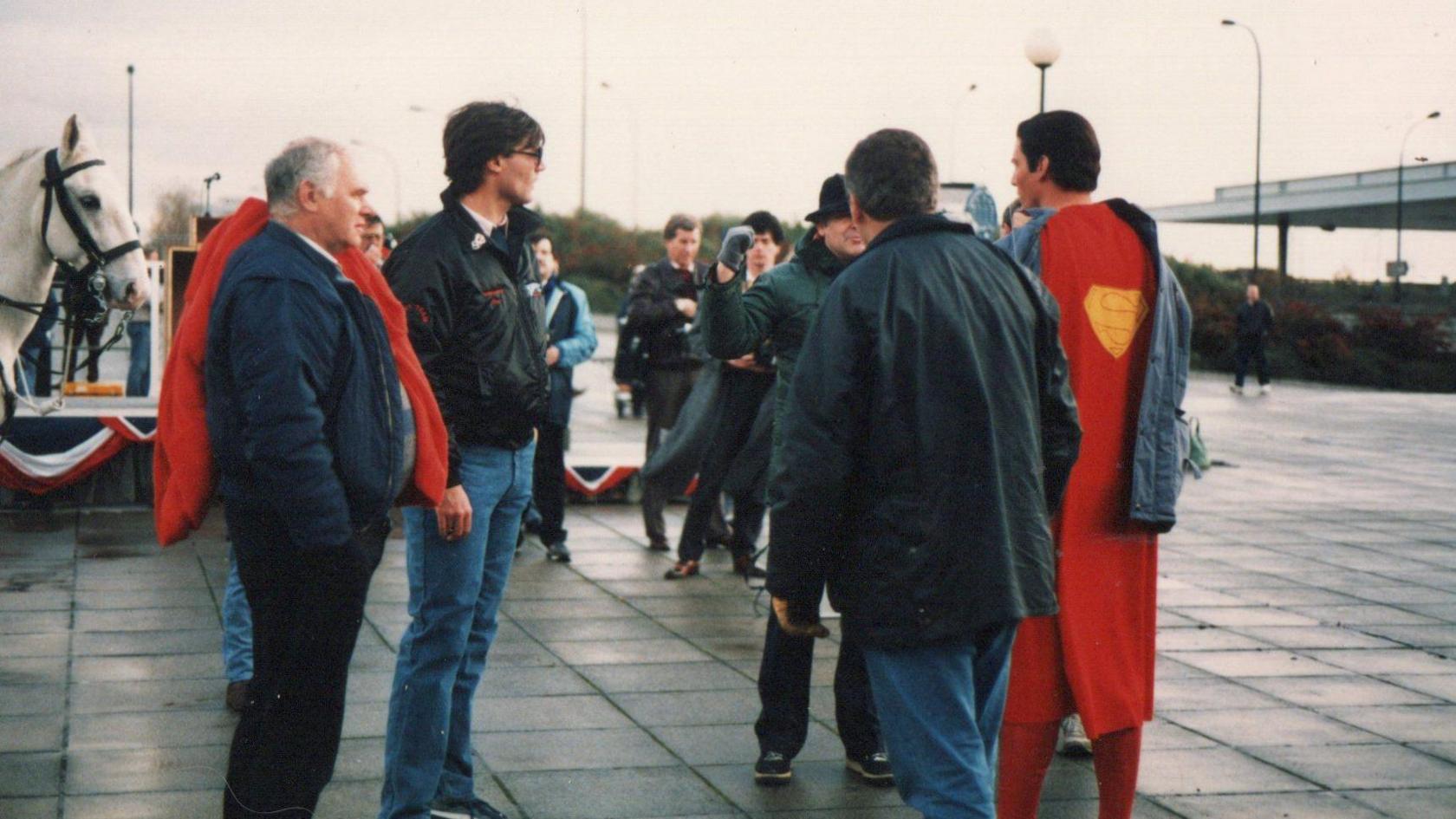 A group of people standing outdoors on a paved area. One person wears a Superman costume with a red cape and a yellow "S" emblem. A white horse is on the left, with streetlights, an overpass and greenery in the background.
