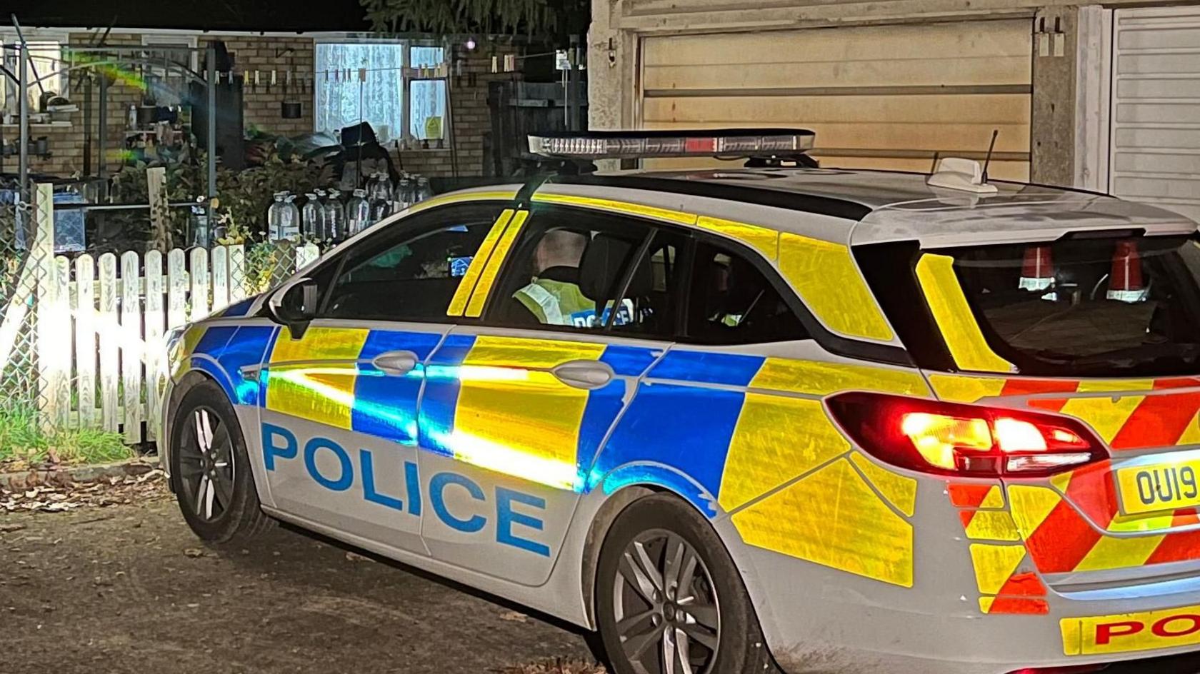 A police car outside a bungalow. It is dark and the car's lights are shining on to a wooden fence and into the garden.
