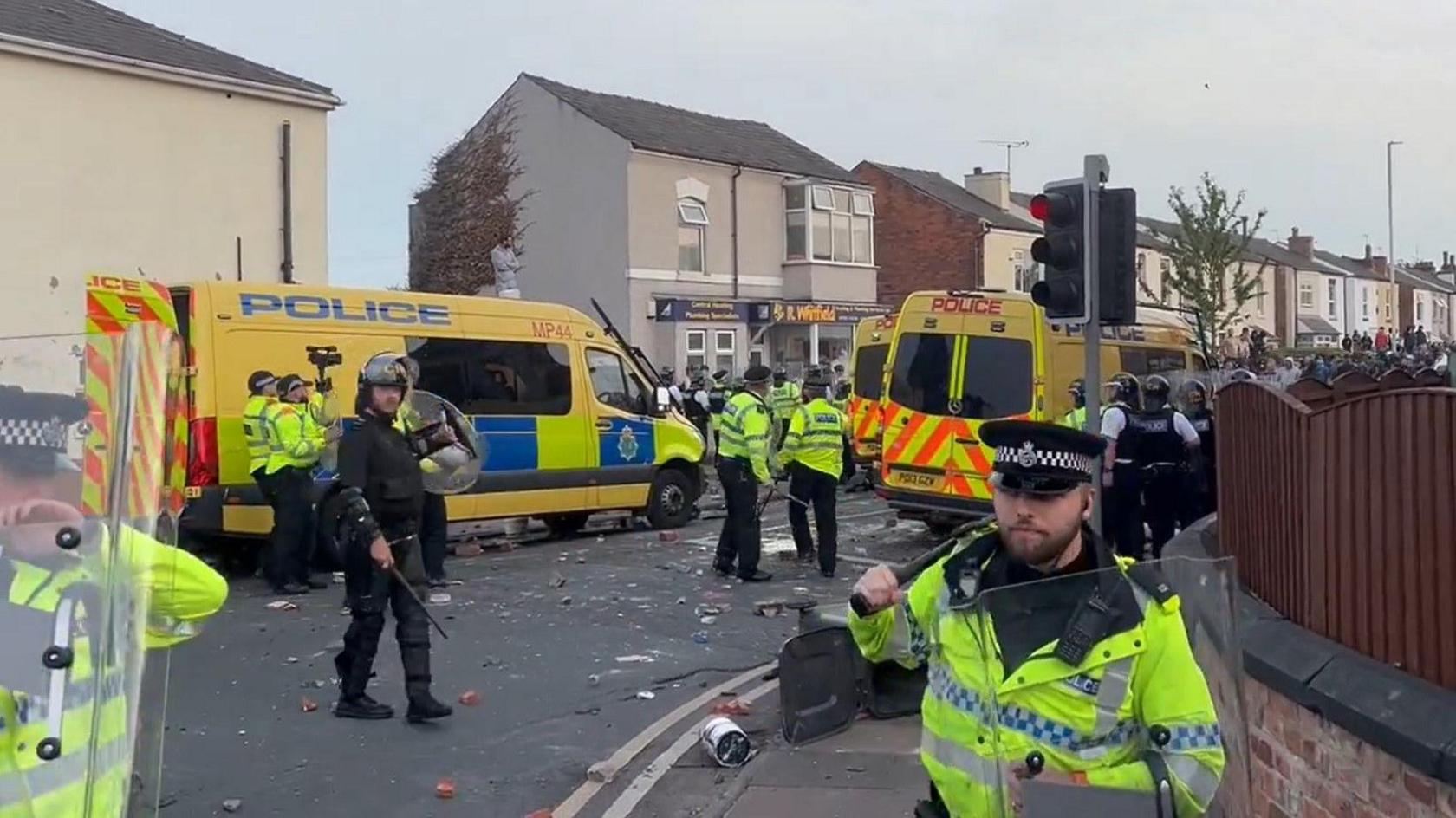 Police officers with riot gear gather on a street corner in Southport, where debris can be seen strewn across the road. Police vehicles also line up.