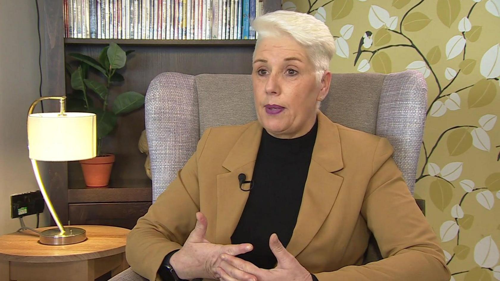 Samantha Hoskins, a woman with short white hair wearing a mustard coloured jacket and black top. She is sitting in an armchair with a table next to her. On the table is a lamp. Behind her is a shelf with dozens of DVDs on it.