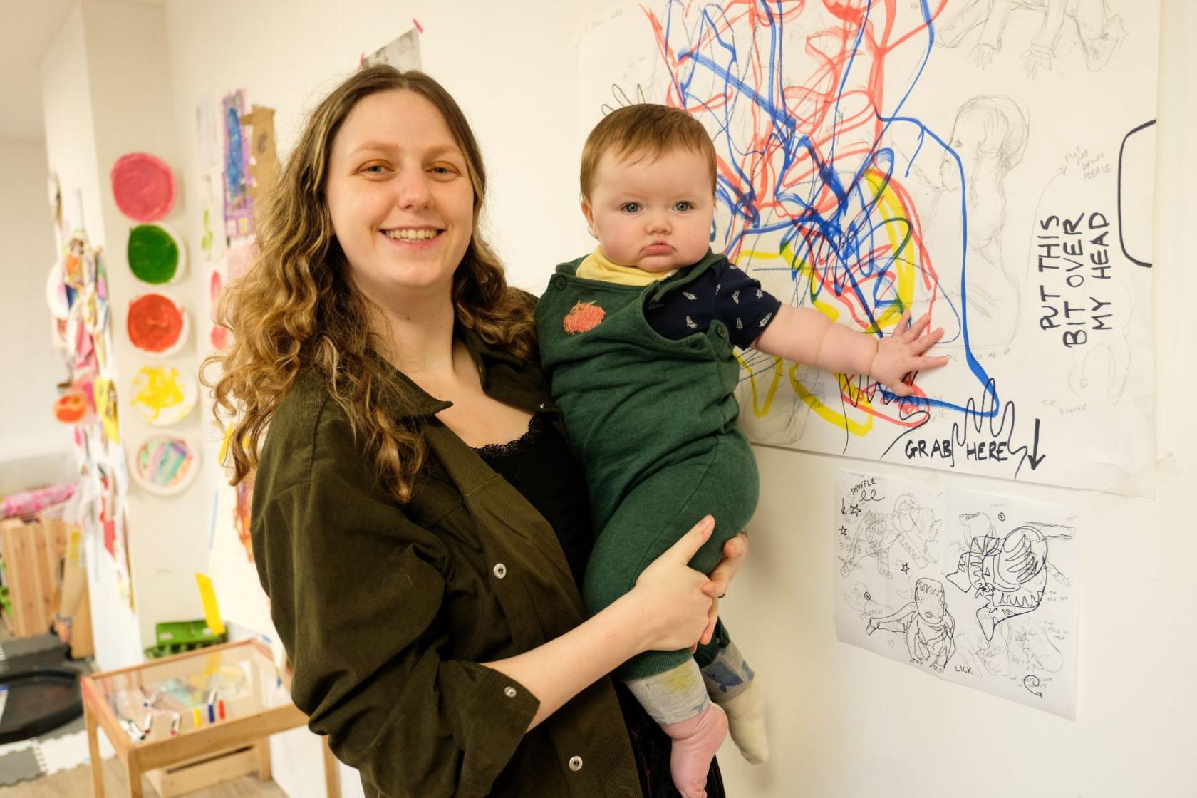 A woman holds a baby and stands in a room that has artworks on the walls. The woman has long wavy brown hair and she is wearing a green shirt. The baby is wearing green dungarees and touching a multi-coloured drawing on the wall.