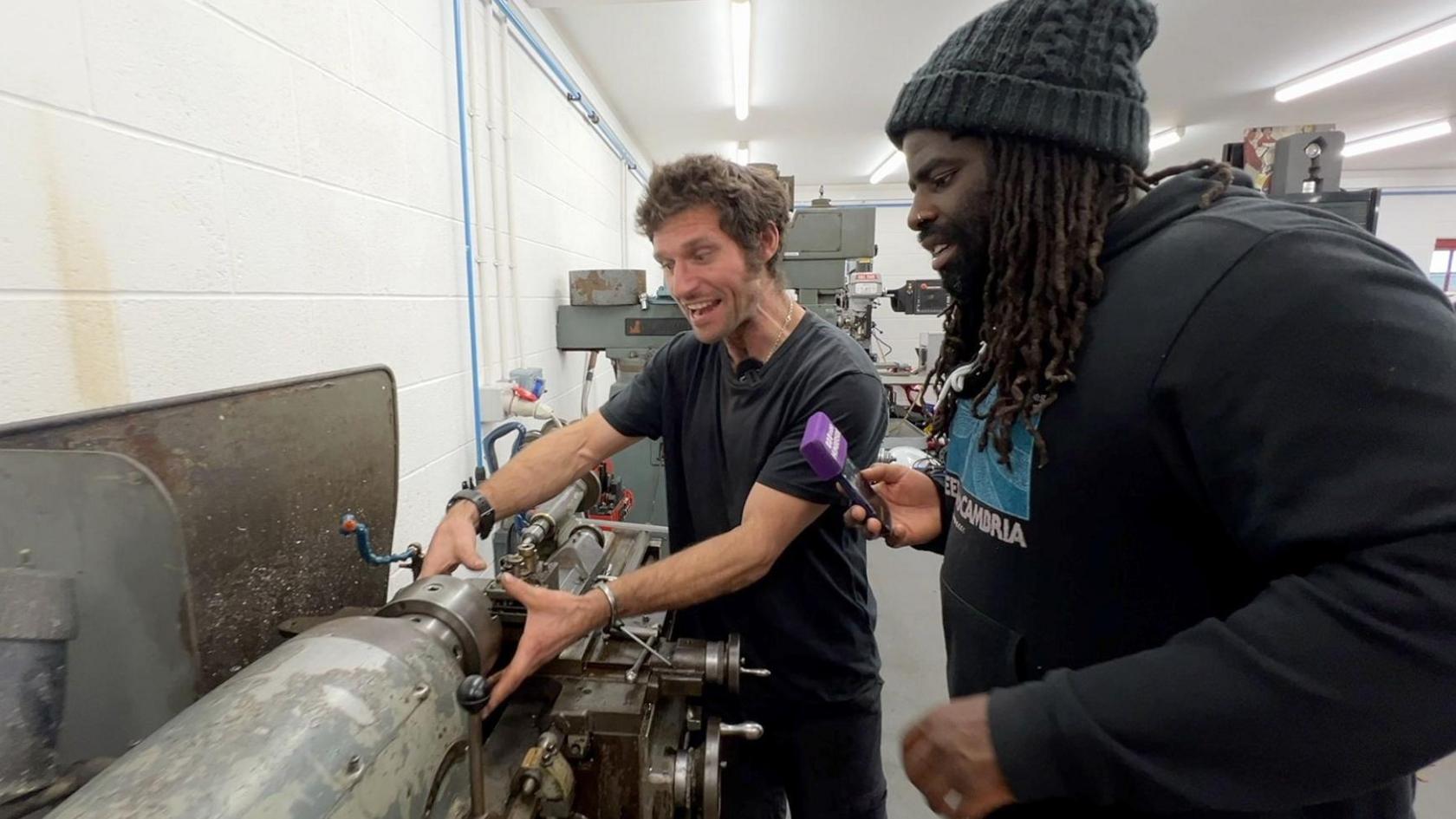 A man with a black T-shirt, short dark hair and sideburns has both hands around a cylindrical machine and is talking to a man on his left, who has dreadlocks and is wearing a black hoodie and a woolly hat. He is holding a phone with a purple microphone cover.