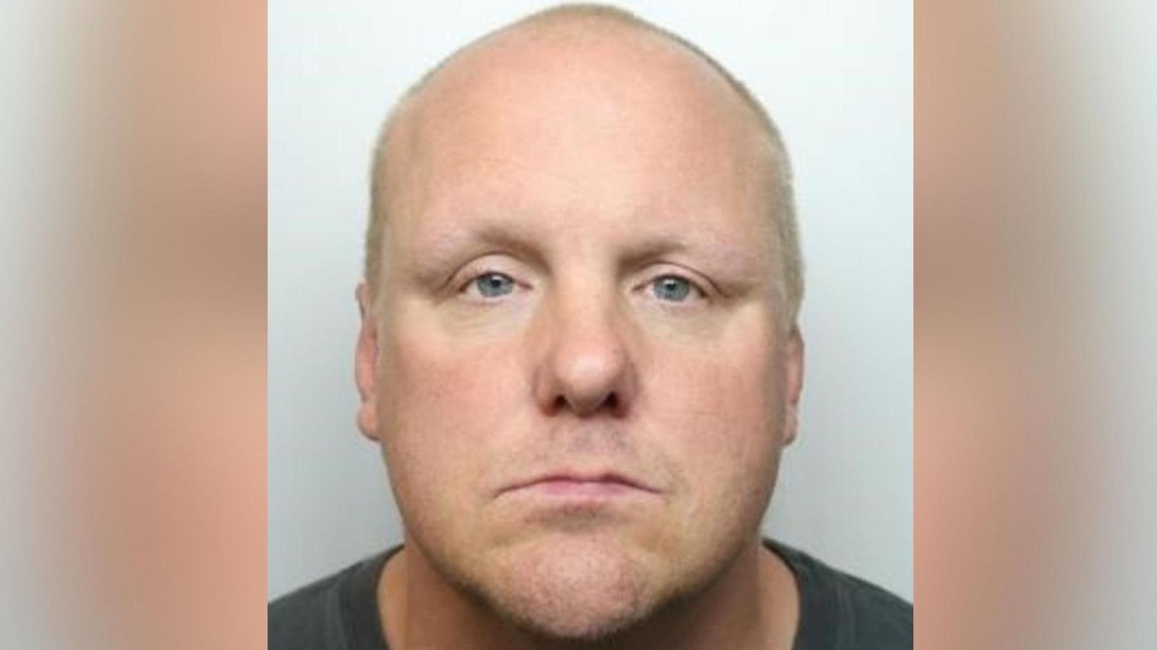 A mugshot of a man who looks directly at the camera. He has no facial hair and his hair is shaved.