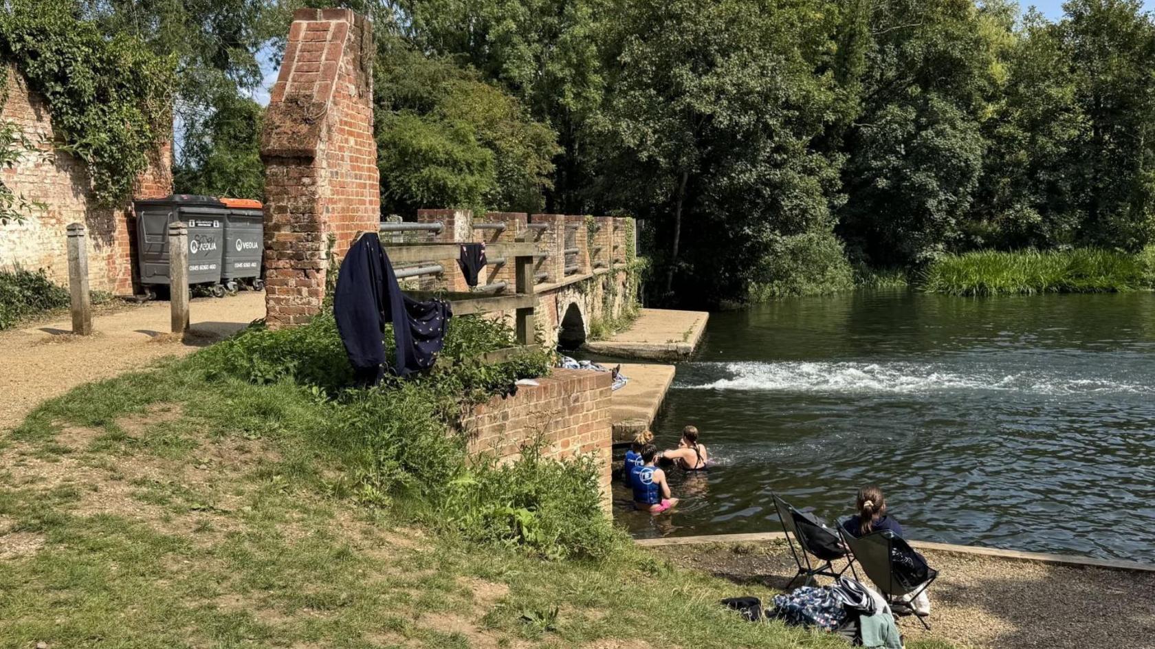 A side view of the brick bridge, with a footpath over the top. Swimmers were in the water, with people sitting on the bank. Trees are in the distance.