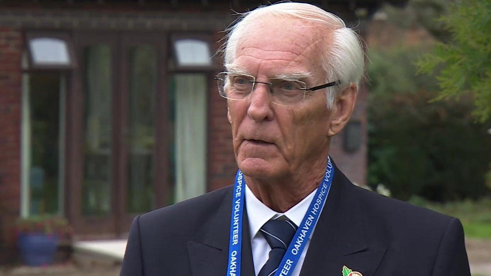 Melville Kendal stands in front of the hospice, which is blurred in the background. He has white short hair, with a side parting, wears clear framed glasses and a dark blue suit, a white shirt and matching dark blue tie with fine white stripes. He is wearing a metal poppy badge and a bright blue lanyard around his neck which says 'Oakhaven Hospice Volunteer'