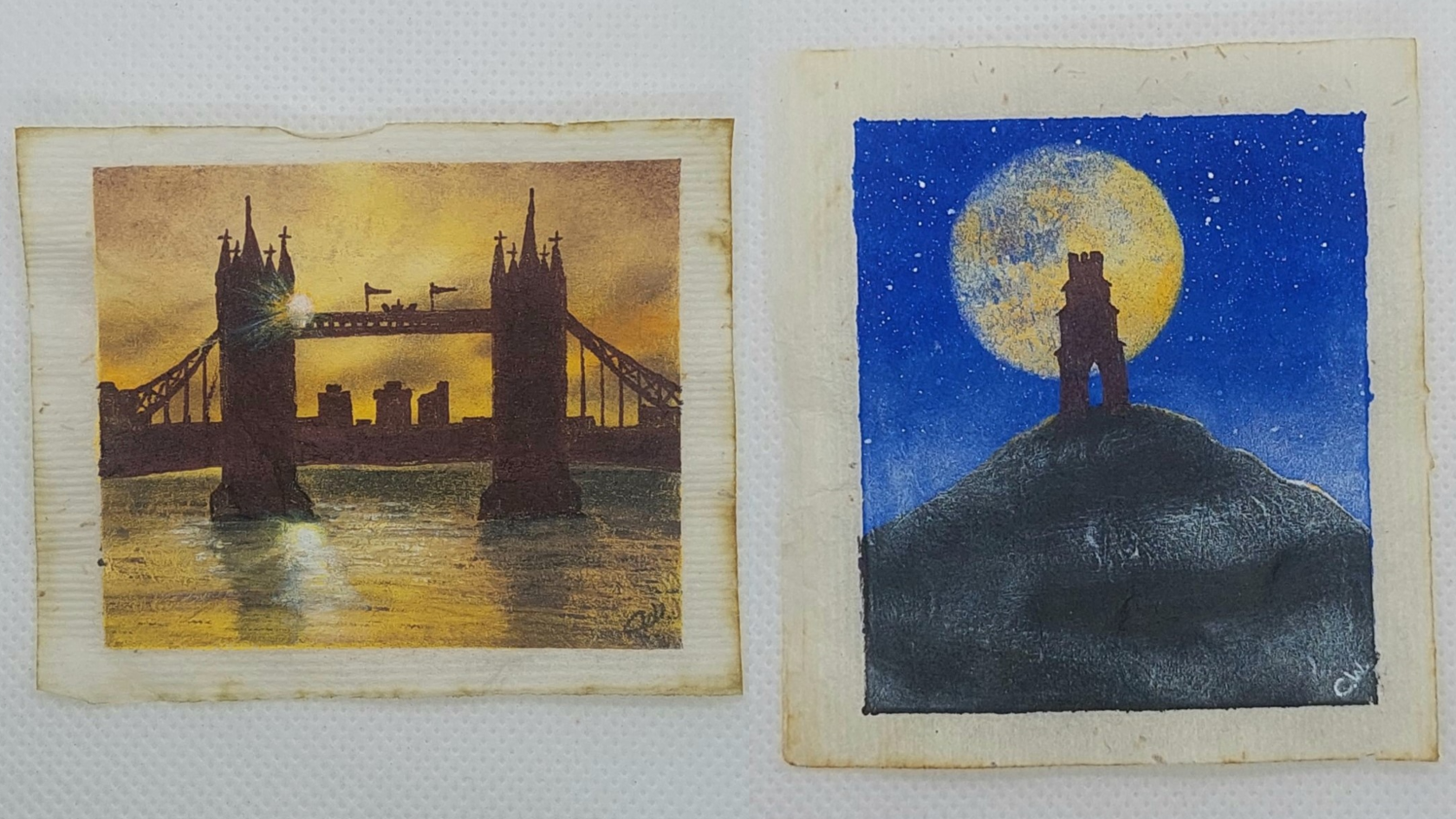 Two teabags - one has Tower Bridge painted on it, whilst the other has Glastonbury Tor on it. Both are in silhouette. Tower Bridge has a golden sky, reflected back in the water below, and Glastonbury Tor stands in front of a golden moon and deep blue sky.