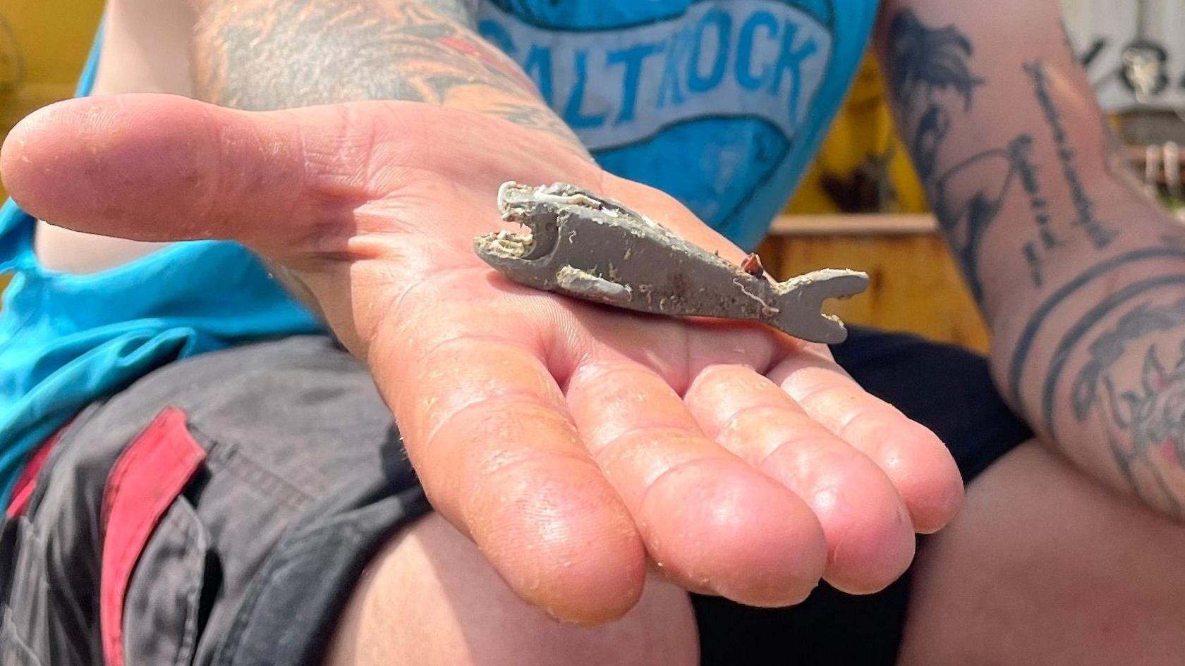 A close-up of Richard West's open hand which has a small, grey Lego shark on it. The piece of Lego is worn with scratches in the plastic. Mr West is sat on a fishing trawler. He is wearing a blue vest top and shorts.