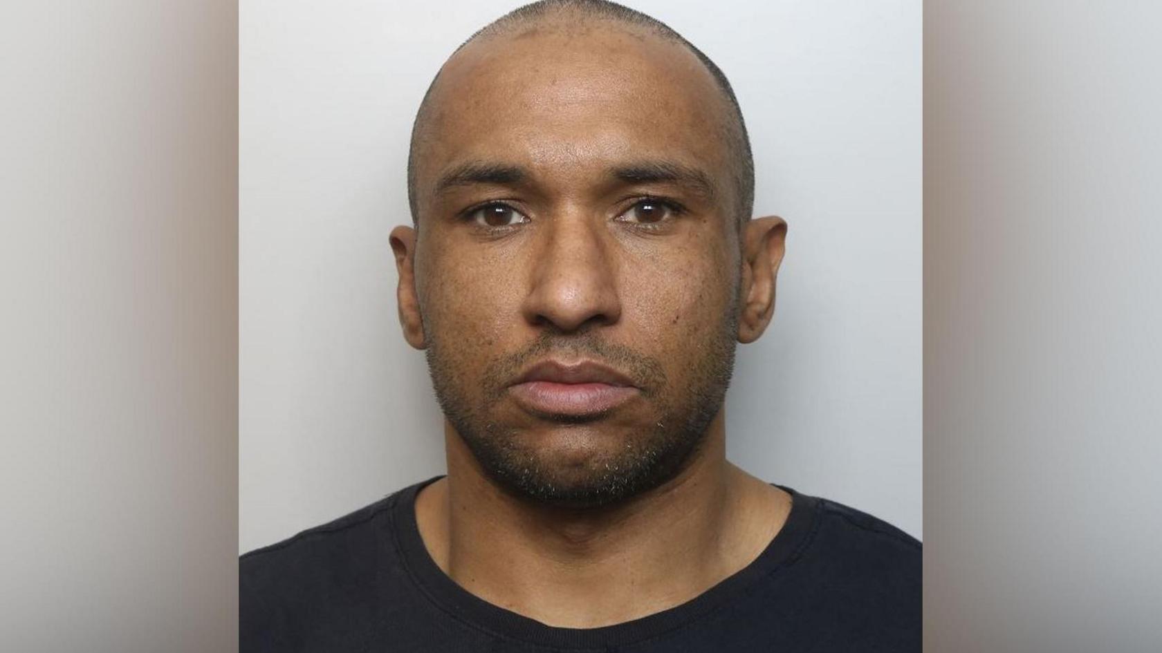 A police custody photo of a man with a shaved head and black top standing against a plain white background.