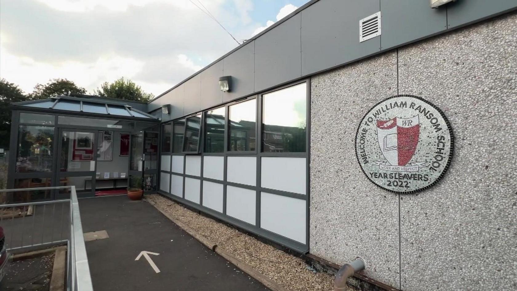 The entrance to William Ransom Primary School. A grey building with a school badge by the glass entrance.