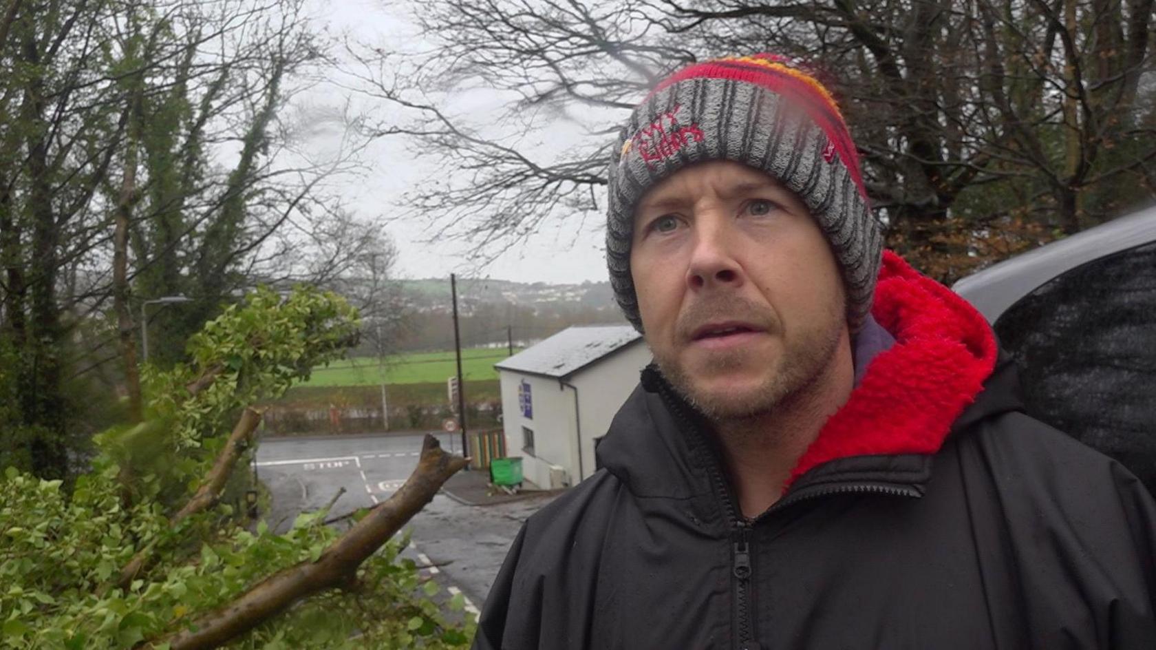 Marc Morris stands by the side of a rural road next to his car. A large tree is down on the verge behind him. 